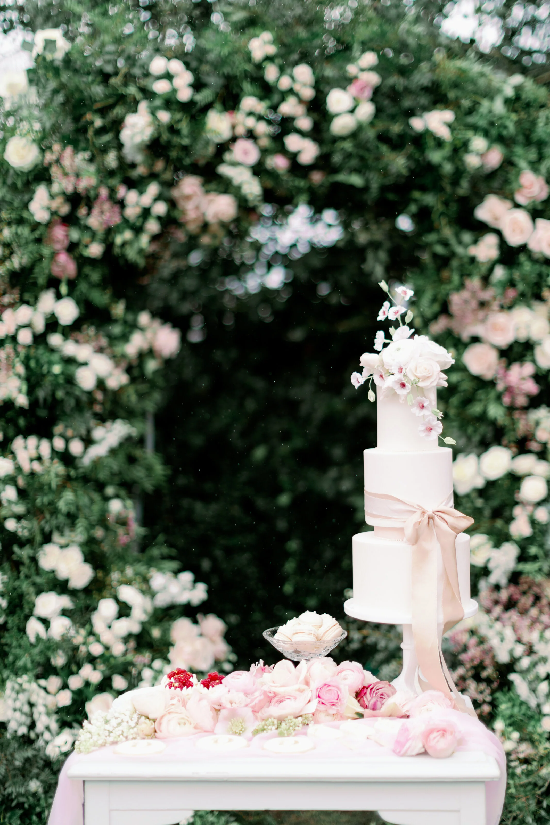 A white three-tier wedding cake decorated with pink and white flowers, tied with a pink ribbon, on a stand in front of a floral arch with pink and white roses.