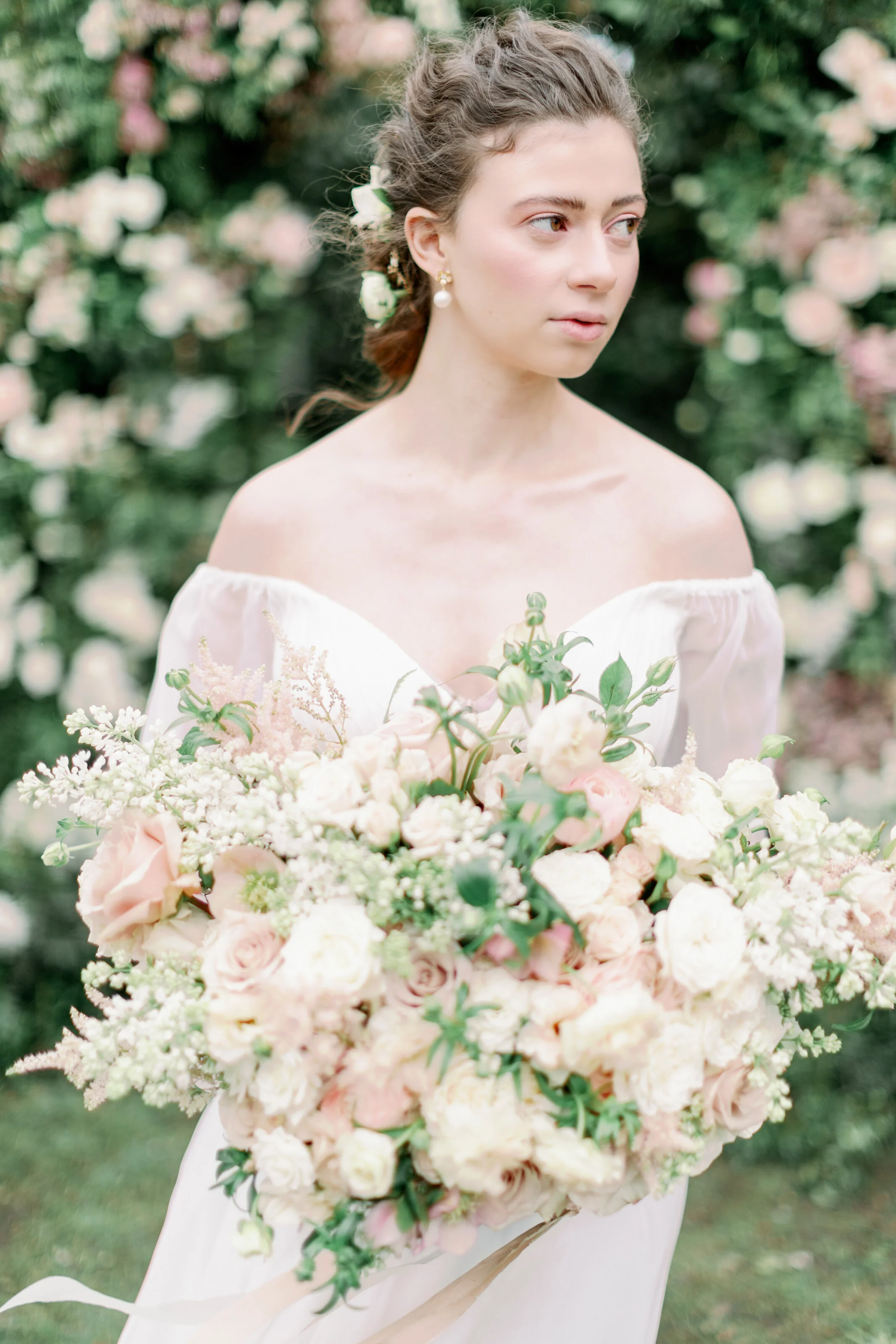 A young woman in a white off-the-shoulder dress holding a large bouquet of pink and white roses with greenery, standing outdoors with blurred pink and white flowers in the background.