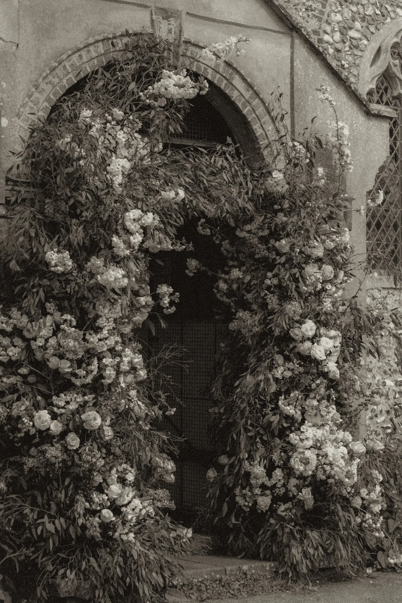 A stone building with an arched window partially covered by dense, flowering bushes with many white blossoms.