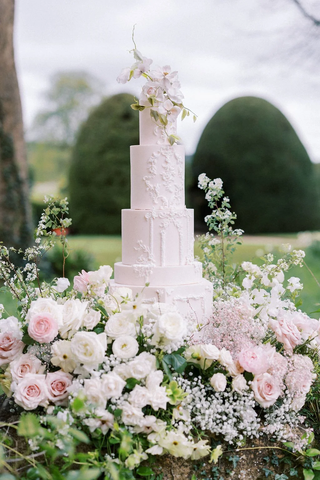 Elegant white wedding cake on a floral display with pink and white roses and greenery outdoors.