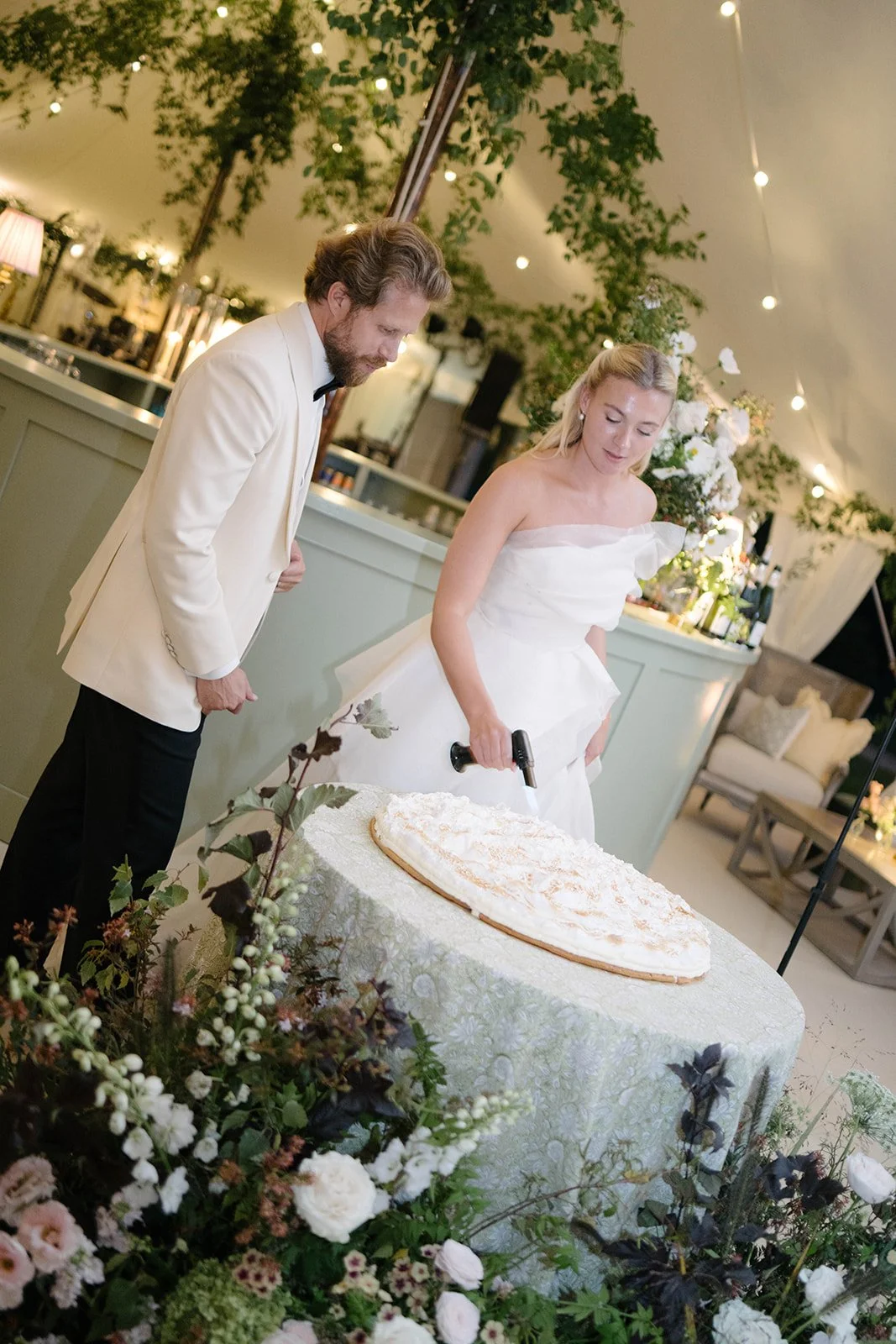 Bride and groom flame torching their lemon meringue pie inside a sailcloth marquee at Cornwell Manor beneath a foliage ceiling installation styled by The Botany House wedding florist