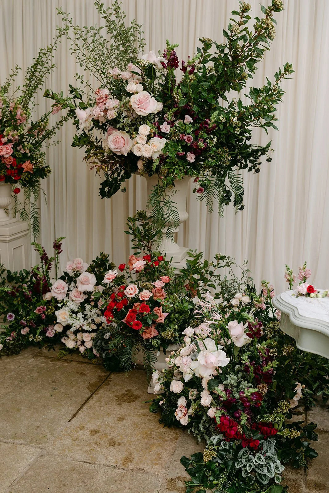 Elegant floral arrangement with pink, red, and white flowers, including roses and greenery, in a white urn on a beige floor with a cream curtain background. Kin House, The Botany House, Get wed