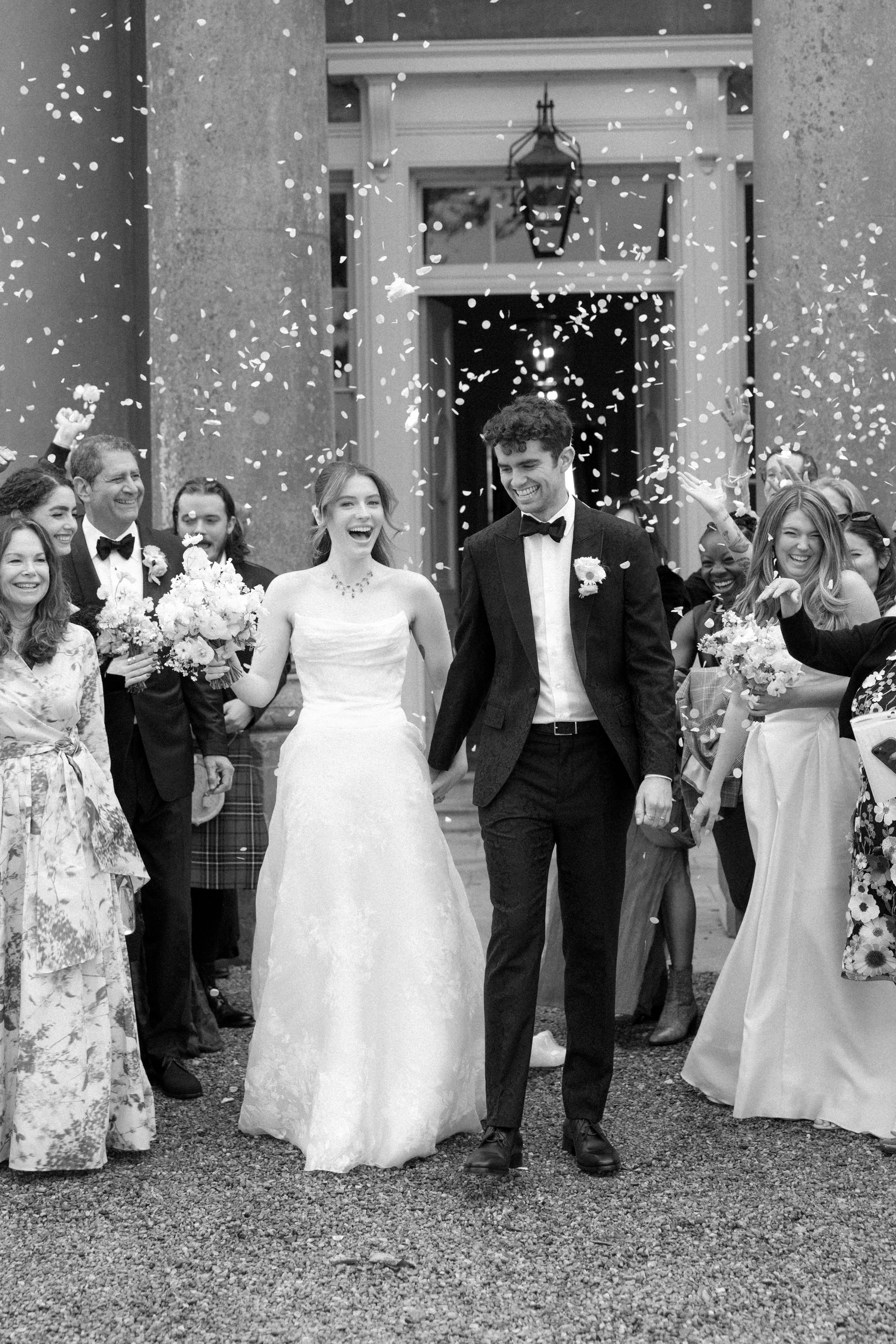 Black and white photo of a wedding celebration showing a bride and groom walking out of a building with guests cheering and throwing confetti.