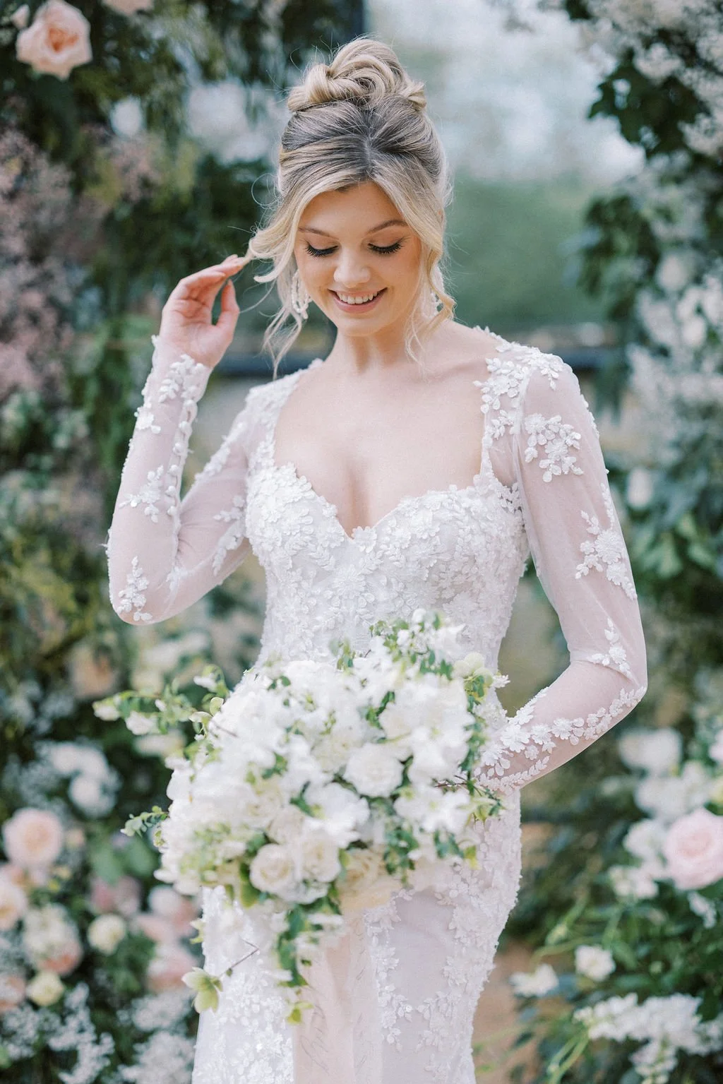 A bride in a long-sleeved, lace wedding dress holding a bouquet of white flowers, smiling with eyes closed outdoors surrounded by greenery and flowers.