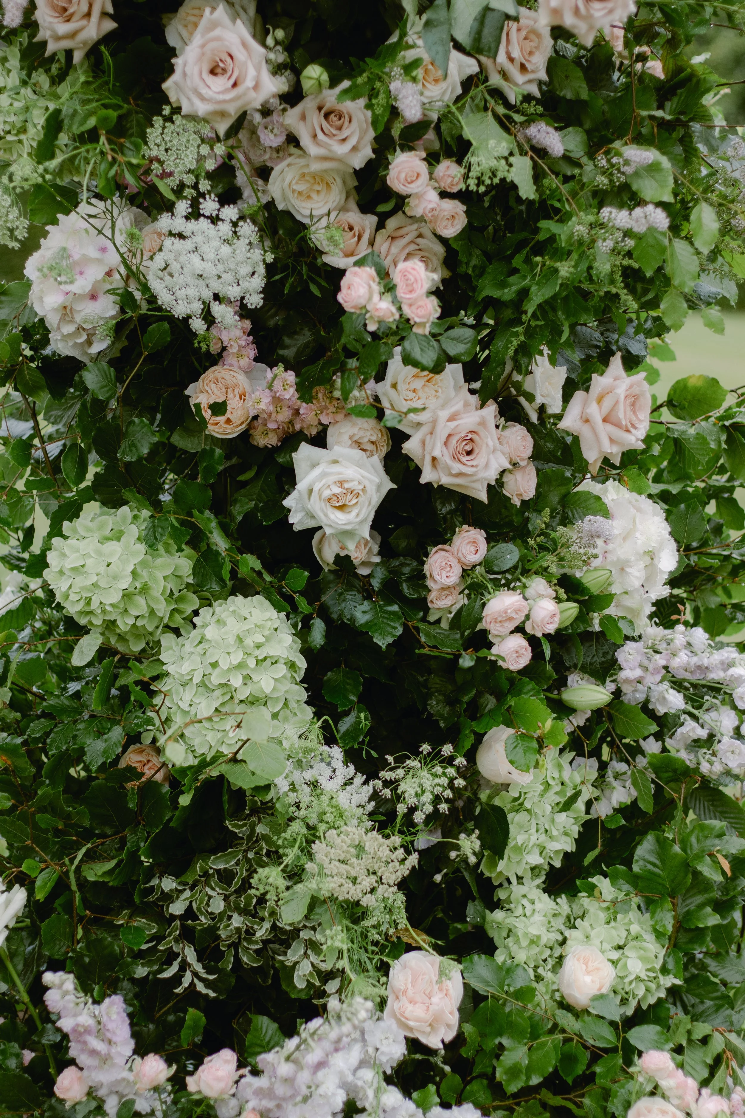 A close up of a floral archway with white and pale pink roses, hydrangeas, and various white flowers with green foliage.