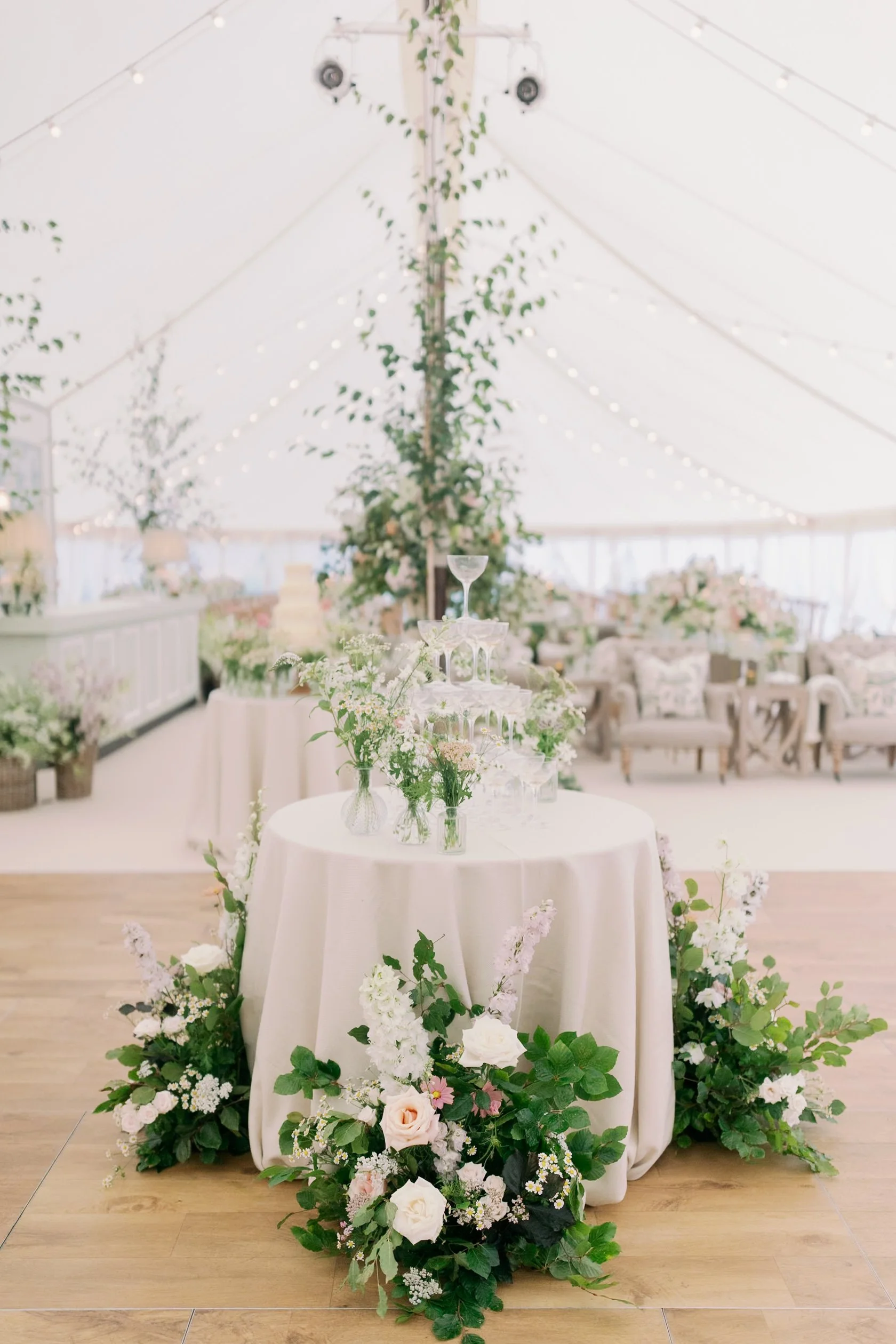 Elegant wedding reception centerpiece with champagne glasses, flowers, and a champagne tower on a round table with a white tablecloth, surrounded by floral arrangements, inside a decorated sailcloth marquee in Norfolk.
