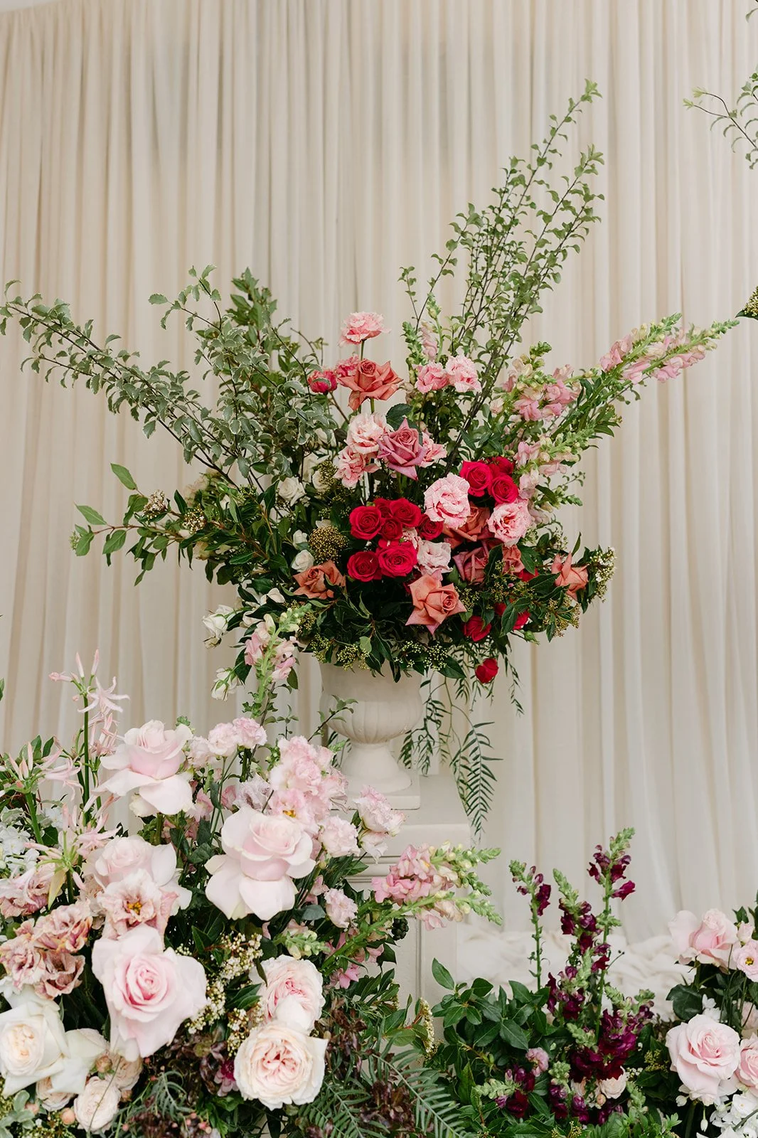 A large arrangement of pink, red, and white flowers with green foliage displayed in a white urn on a pedestal against a light-colored curtain background. Wedding Ceremony Kin House The Botany House