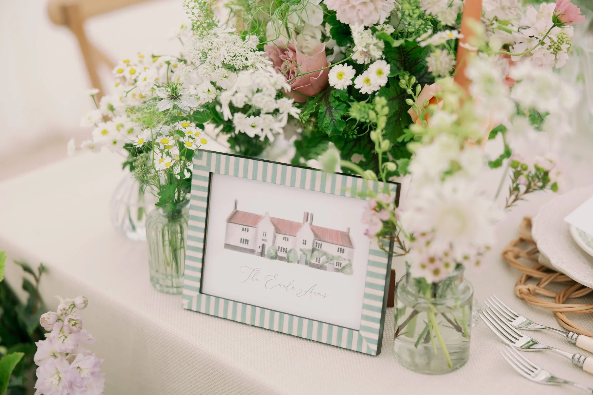 Table centerpiece with budvases , a framed watercolor illustration of a house with the words 'The Earl Arms,' and silver forks on a beige tablecloth.