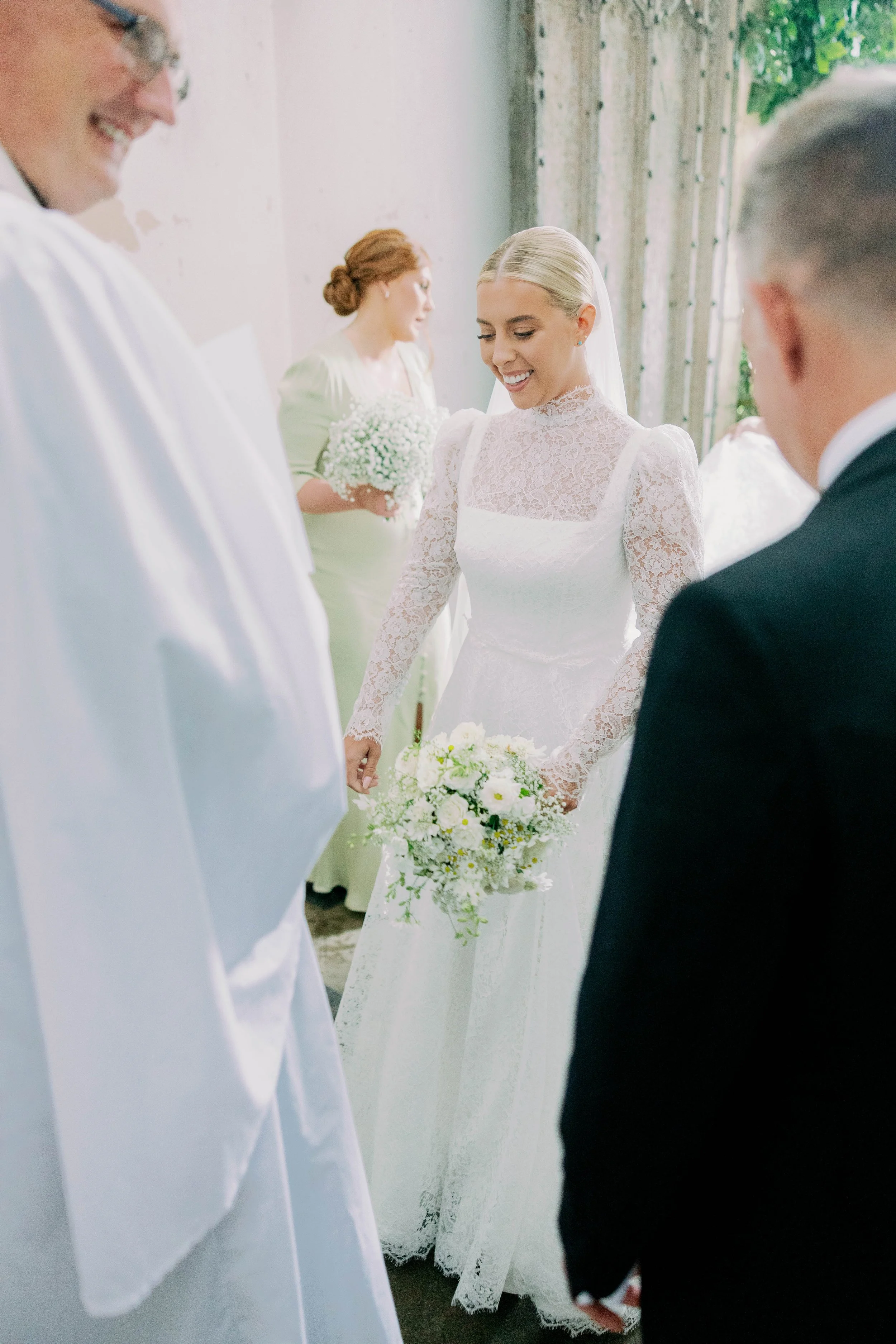 Bride holding a bouquet of white flowers walking into her wedding ceremony surrounded by the vicar and wedding party members.