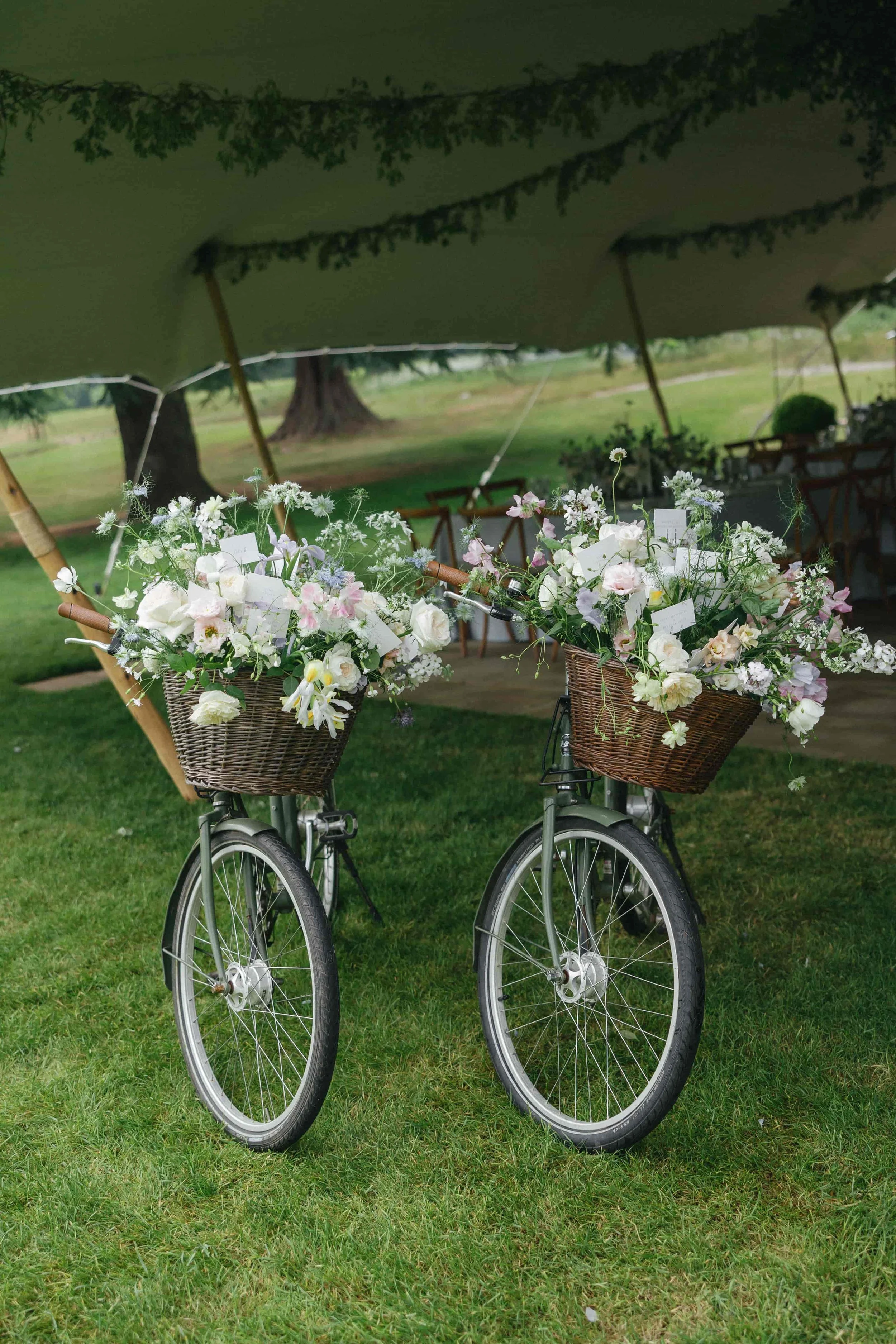 Two vintage bicycles with wicker baskets filled with white and pastel flowers, standing on a grassy lawn under a green canopy with trees and a seating area in the background. The Botany House Florist at Wilderness Reserve, Planned by Attabara