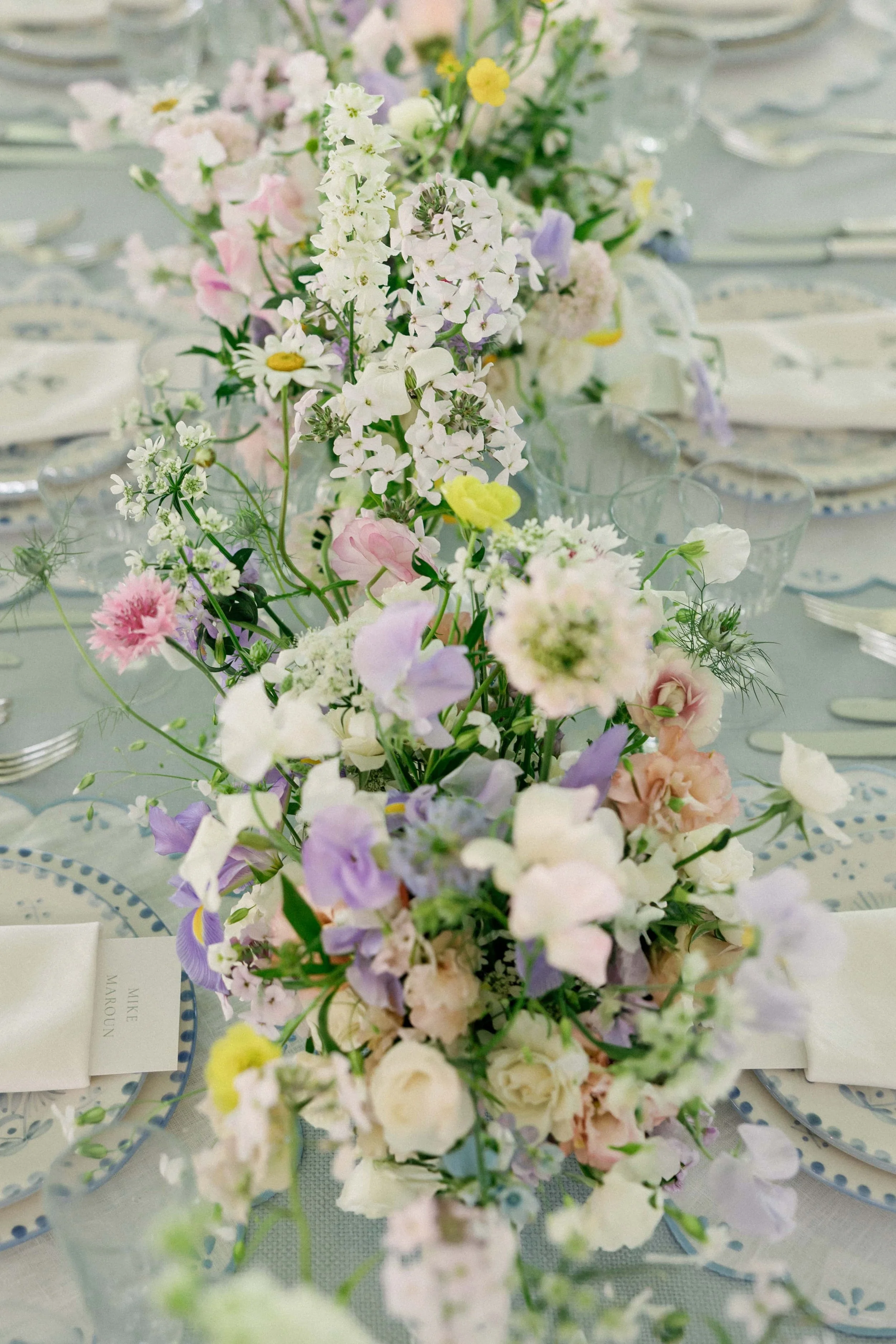 A floral centerpieces with pastel-colored flowers like white, pink, purple, yellow, and green, arranged on a dining table with place settings. Wilderness Reserve, Suffolk Florals By The Botany House