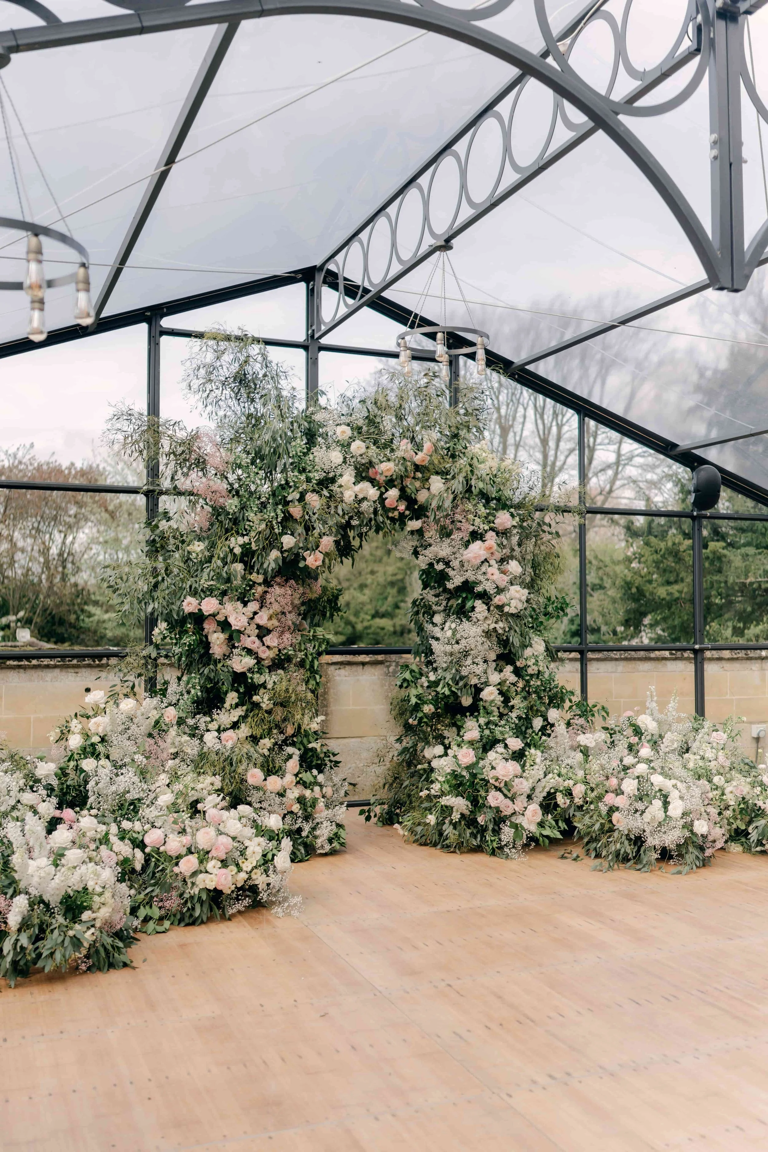 A floral wedding arch decorated with pink and white flowers and greenery, situated inside a glass greenhouse or conservatory with wooden flooring. Sudeley Castle The Cotswolds, Florals by The Botany House