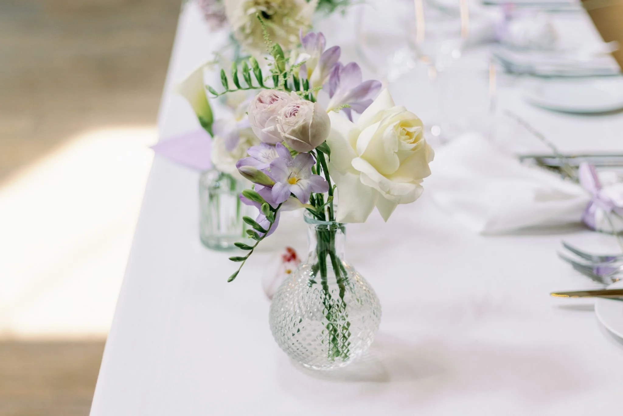 A small glass vase with a textured pattern holding a floral arrangement of white roses, light purple flowers, and green greenery on a white table.