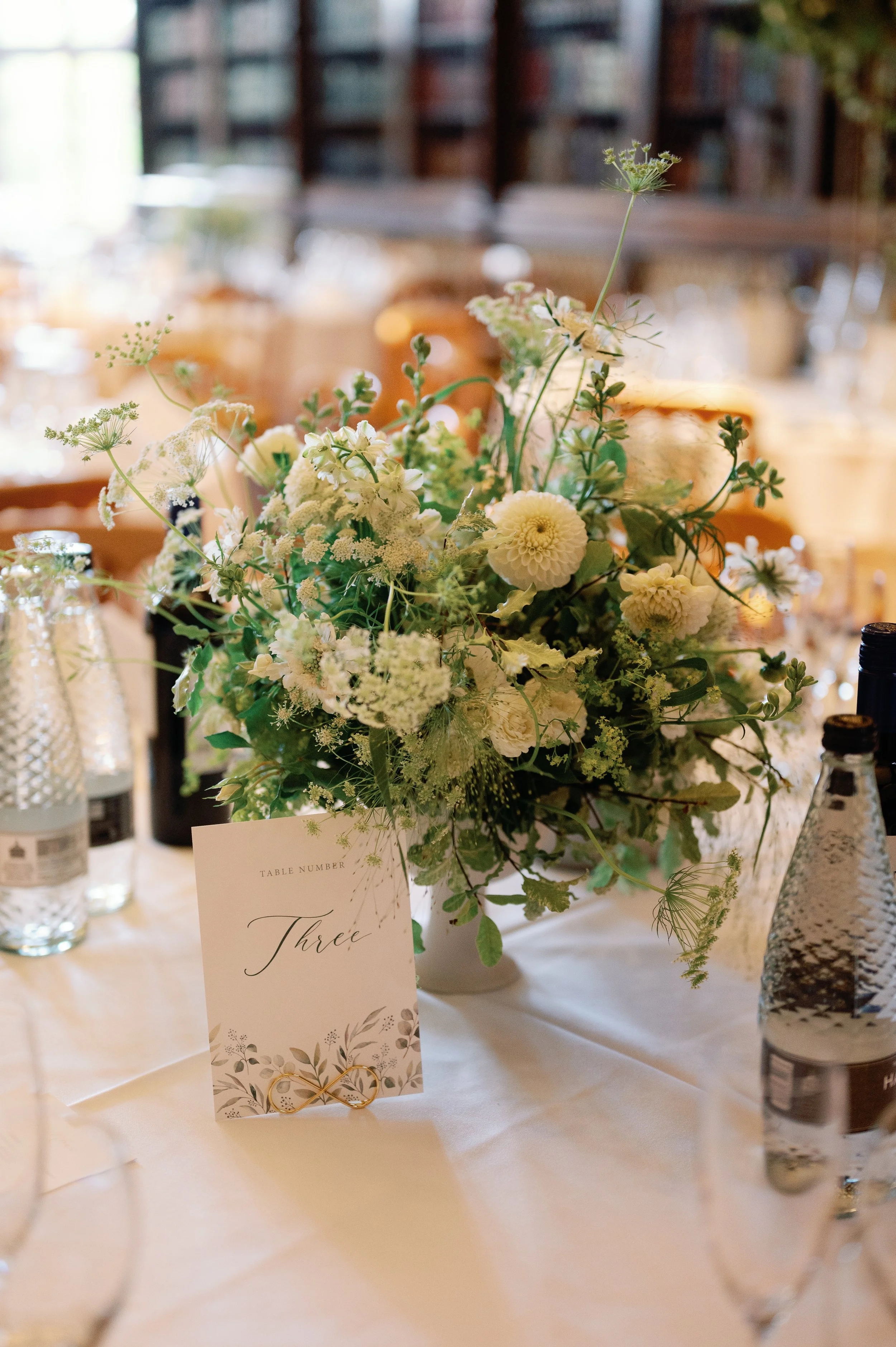 A floral centerpiece on a table with a card labeled "Three" and wedding rings underneath. Bottled water and glassware are also visible.