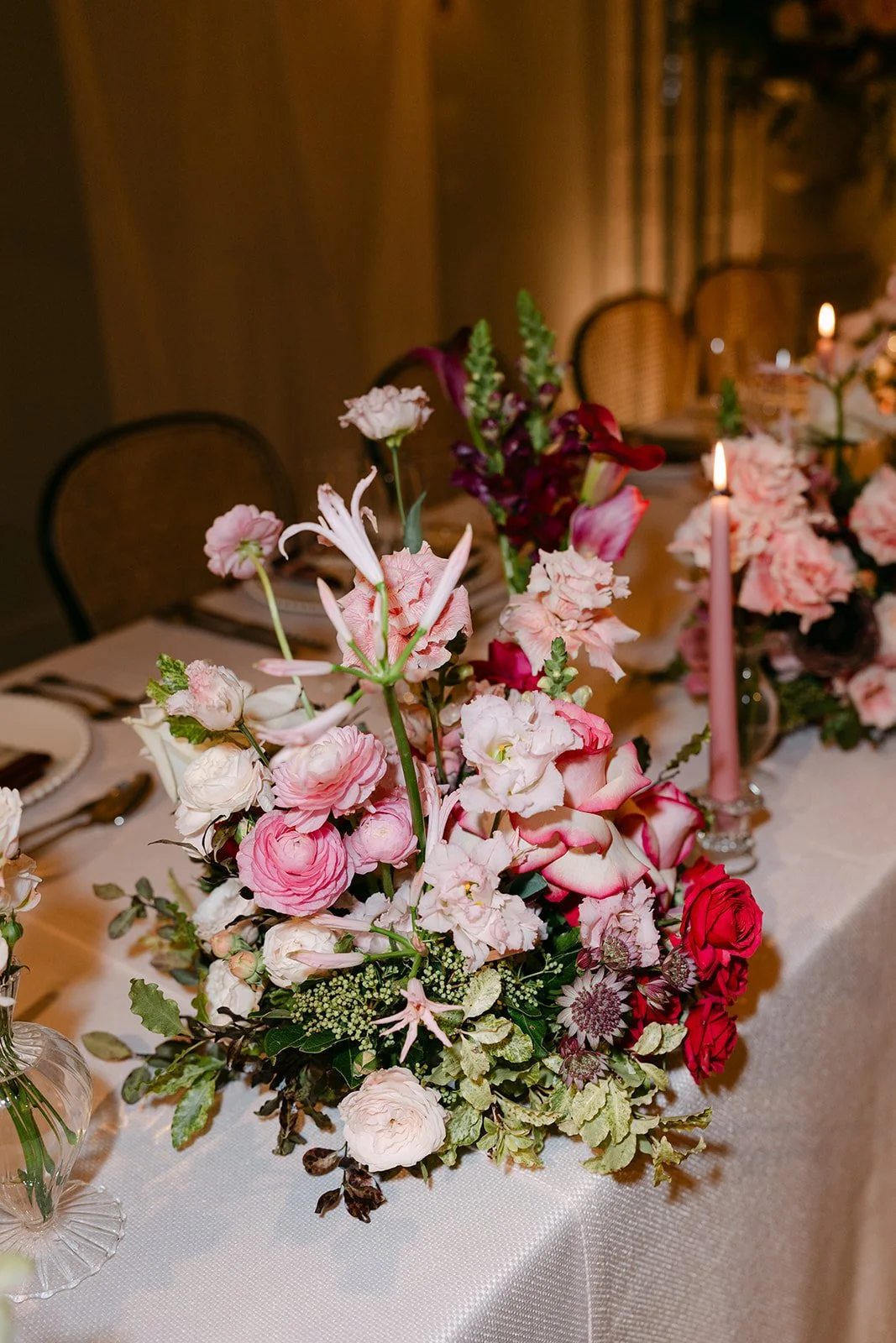 Elegant floral centerpiece on a dining table with pink, white, and deep red flowers, surrounded by candles, set for a formal event. The Botany House Wedding Florist, Kin House The Cotswolds. Planned by Get Wed
