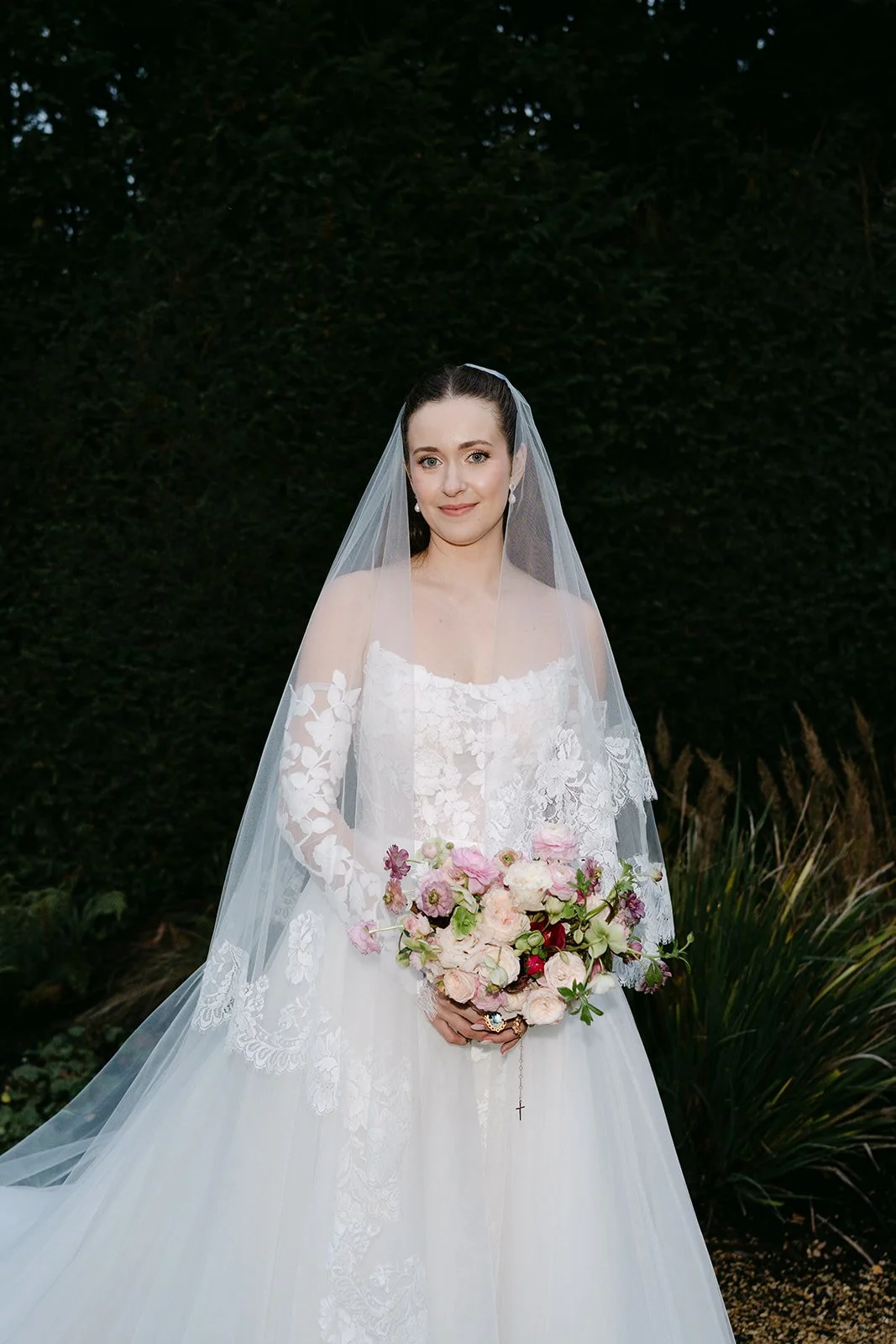 Bride in a white wedding dress holding a bouquet of pink and white flowers, standing outdoors with dark green foliage in the background. The Botany House Wedding Florist, Kin House The Cotswolds. Planned by Get Wed