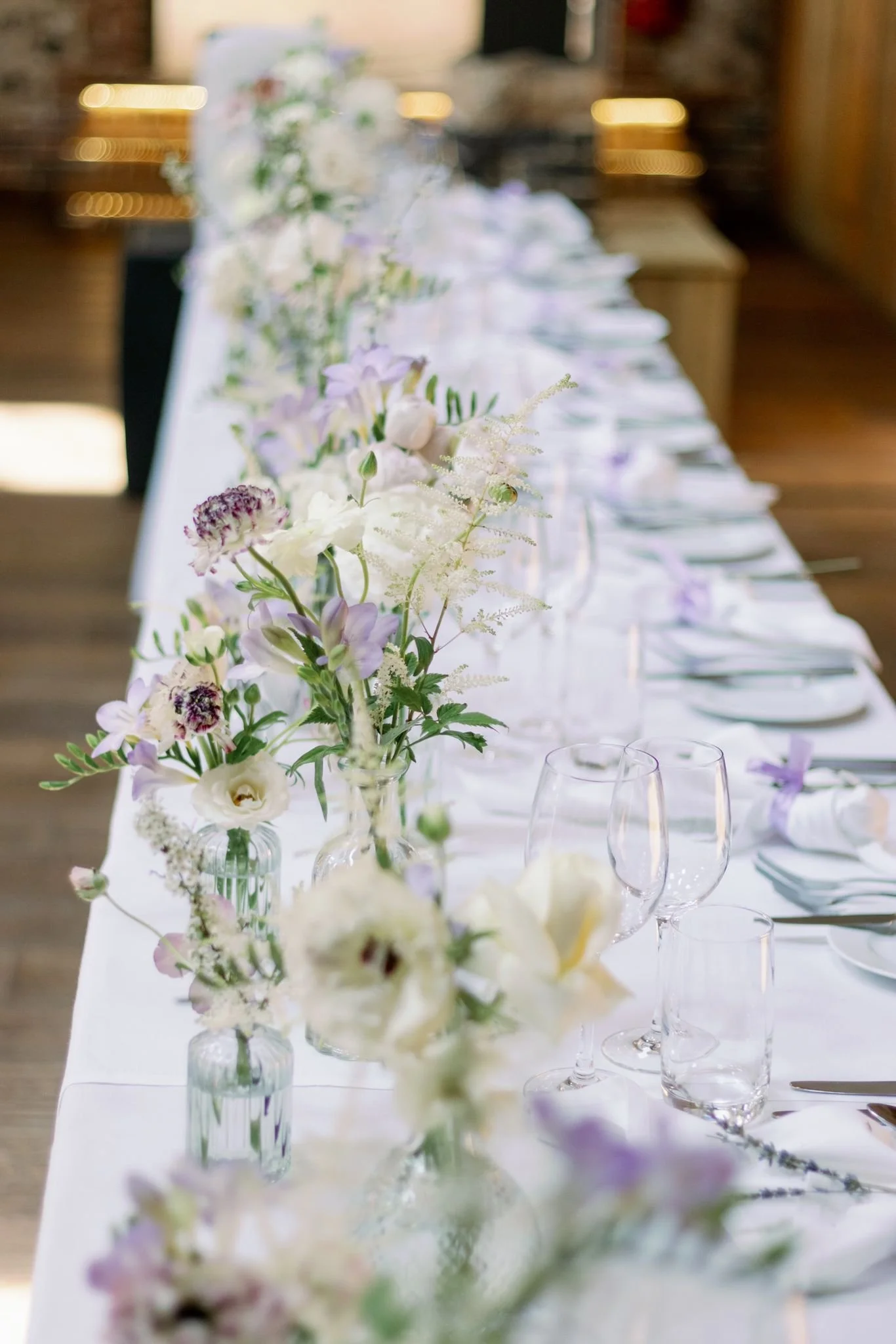 Elegant wedding reception table with white tablecloth and tall floral centerpieces in glass vases, set with wine glasses, water glasses, and silverware, featuring soft purple and white flowers.