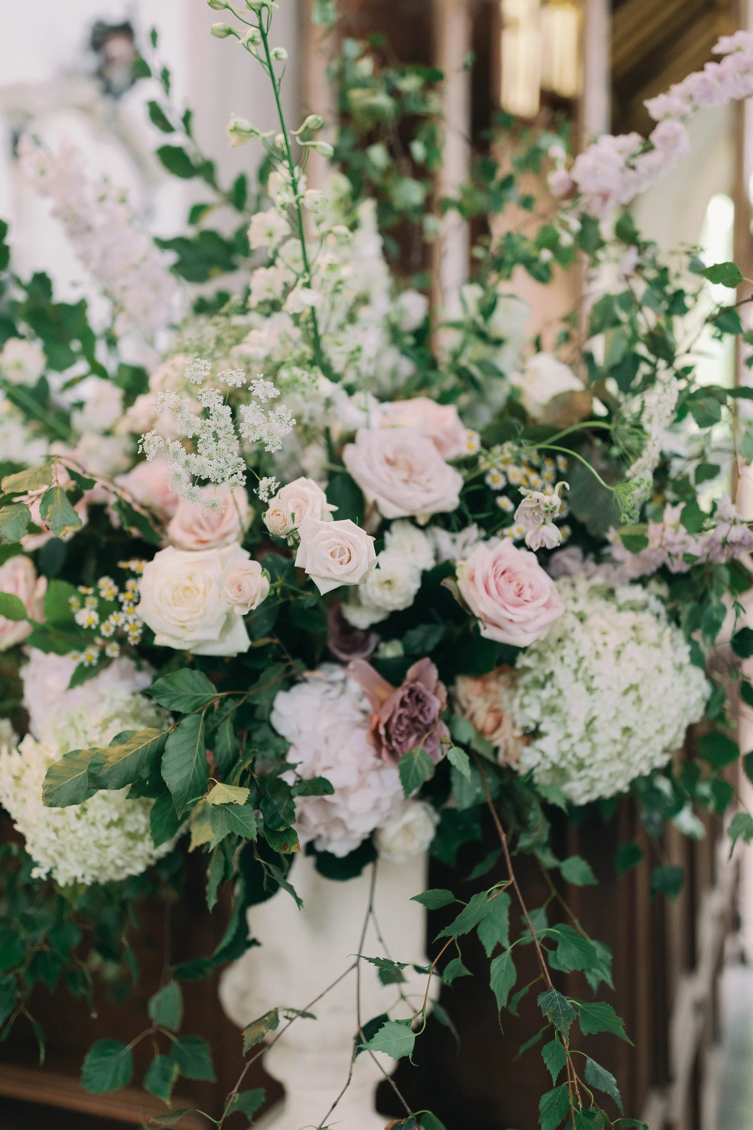 A large urn arrangement of white and pale pink roses, hydrangeas, and various greenery in a white vase, set in a village church in the norfolk countryside