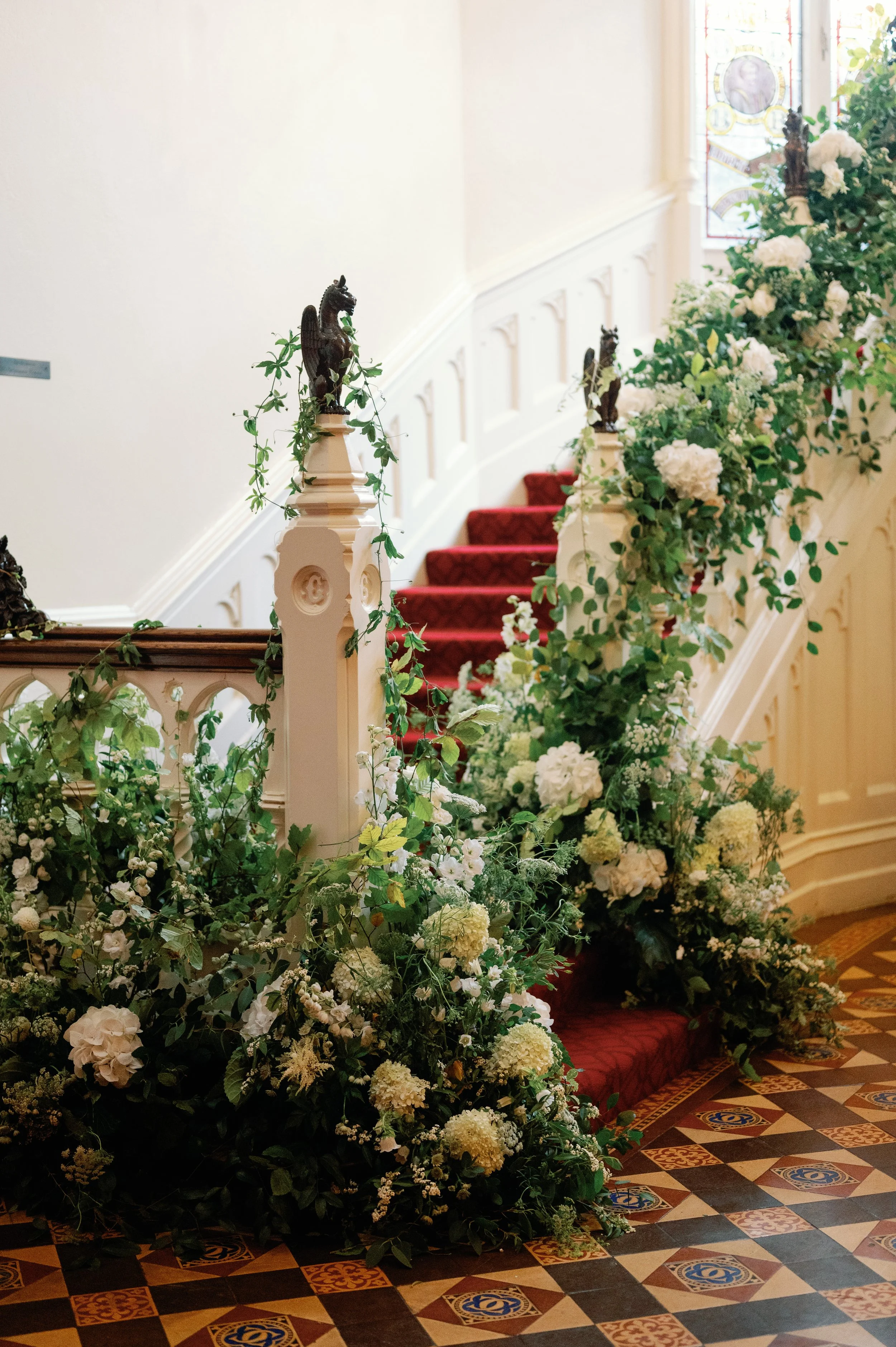 Decorated staircase with white and green floral arrangements, red carpeted steps, and ornate wood and tile flooring in a historic or formal building.