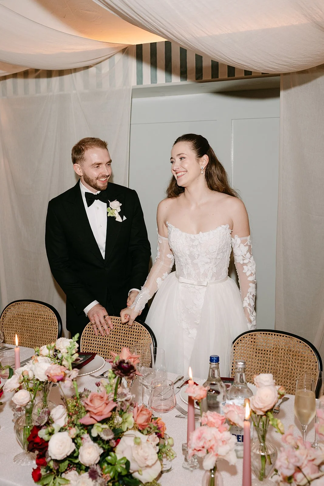 Bride and groom holding hands at their wedding reception, smiling and looking at each other. The bride is wearing an off-shoulder lace wedding dress, and the groom is in a black tuxedo with a bow tie. The table is decorated with pink and white flower