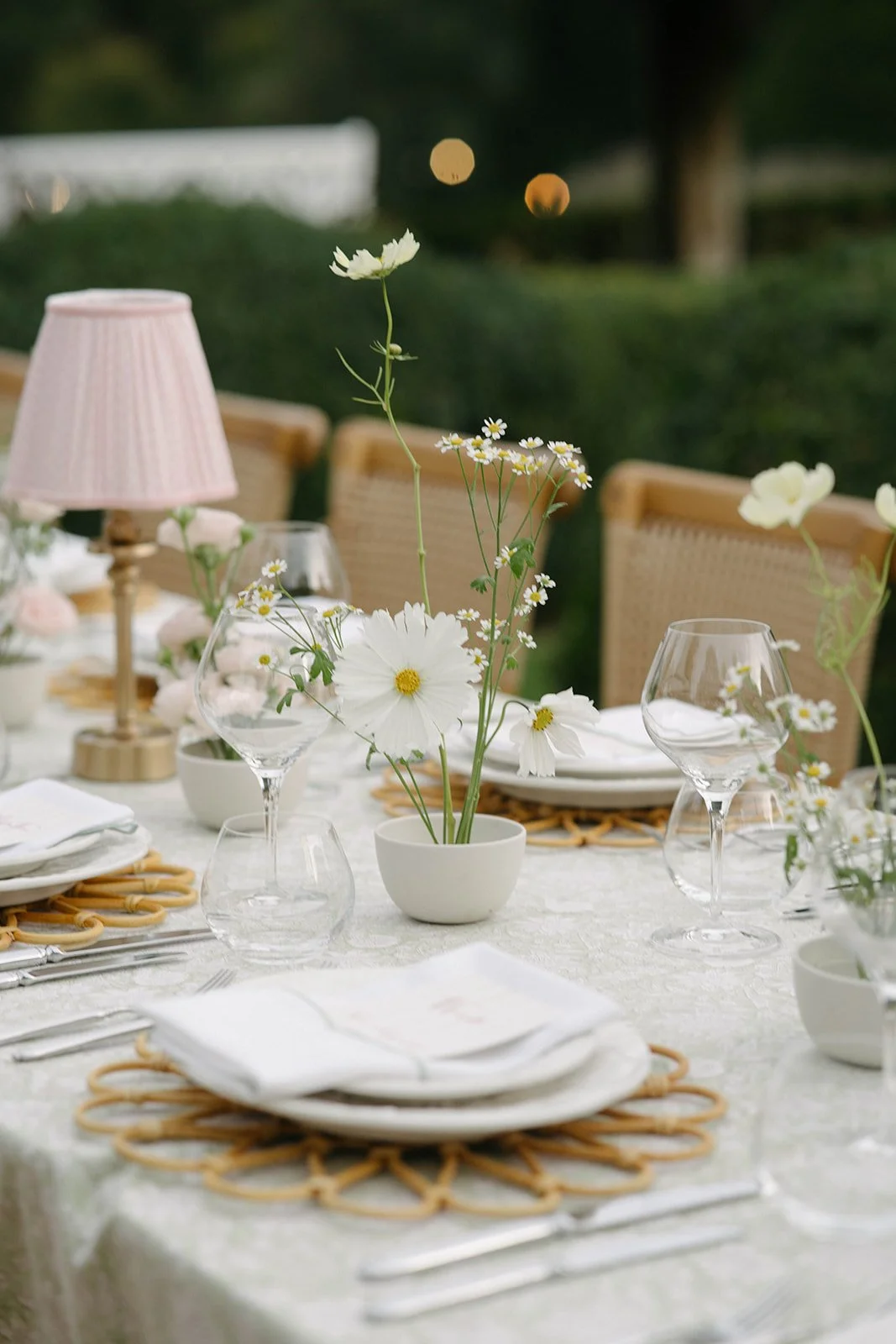 Dining table decorated with white flowers in a white vase, place settings, glasses, and a pink lamp in an outdoor setting - luxury garden-style wedding flowers by The Botany House, wedding florist for Cornwell Manor, Oxfordshire