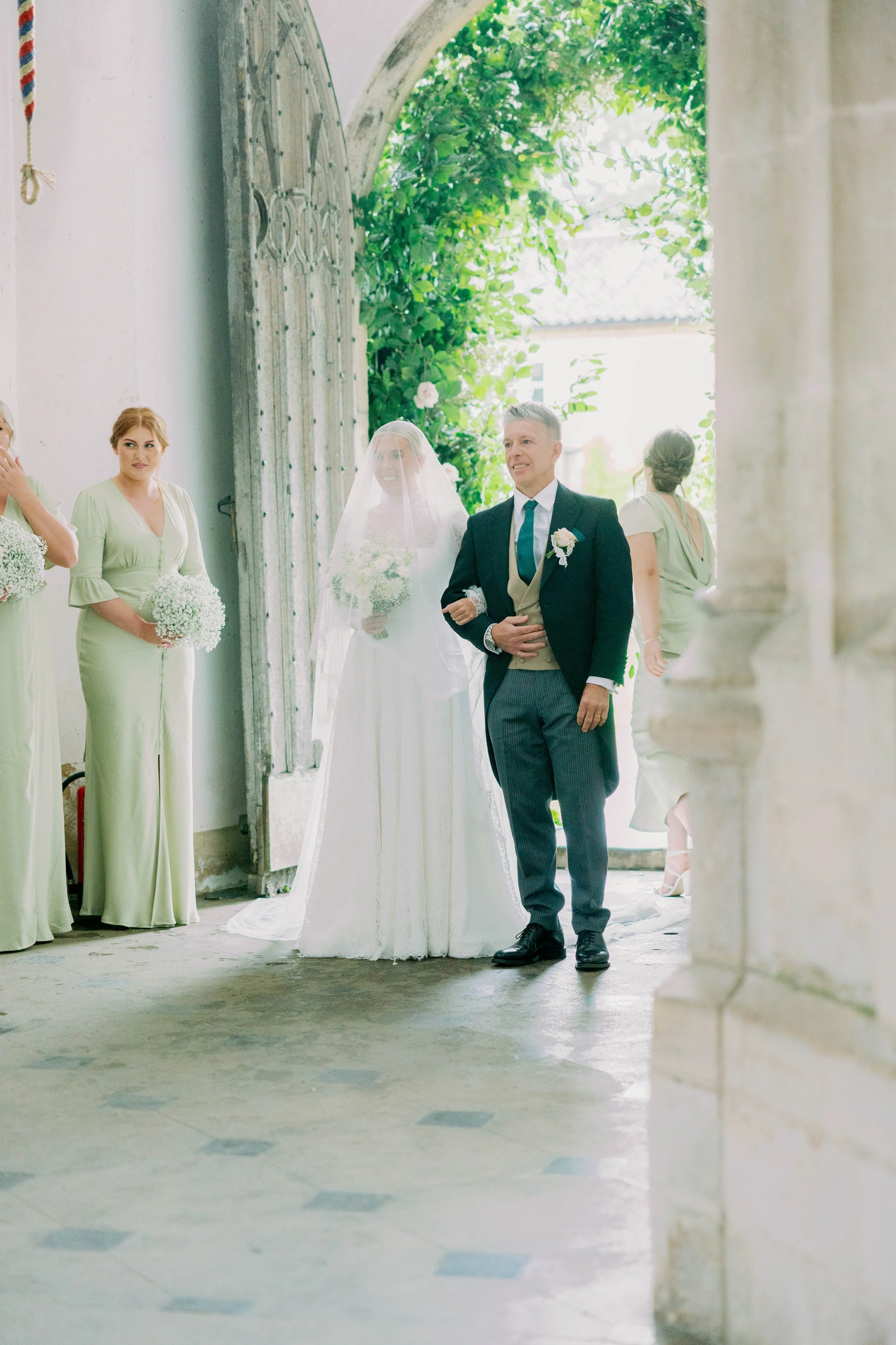 Bride and her father standing at entrance of the church about to walk down the aisle inside a church with bridesmaids and greenery visible outside in the background.