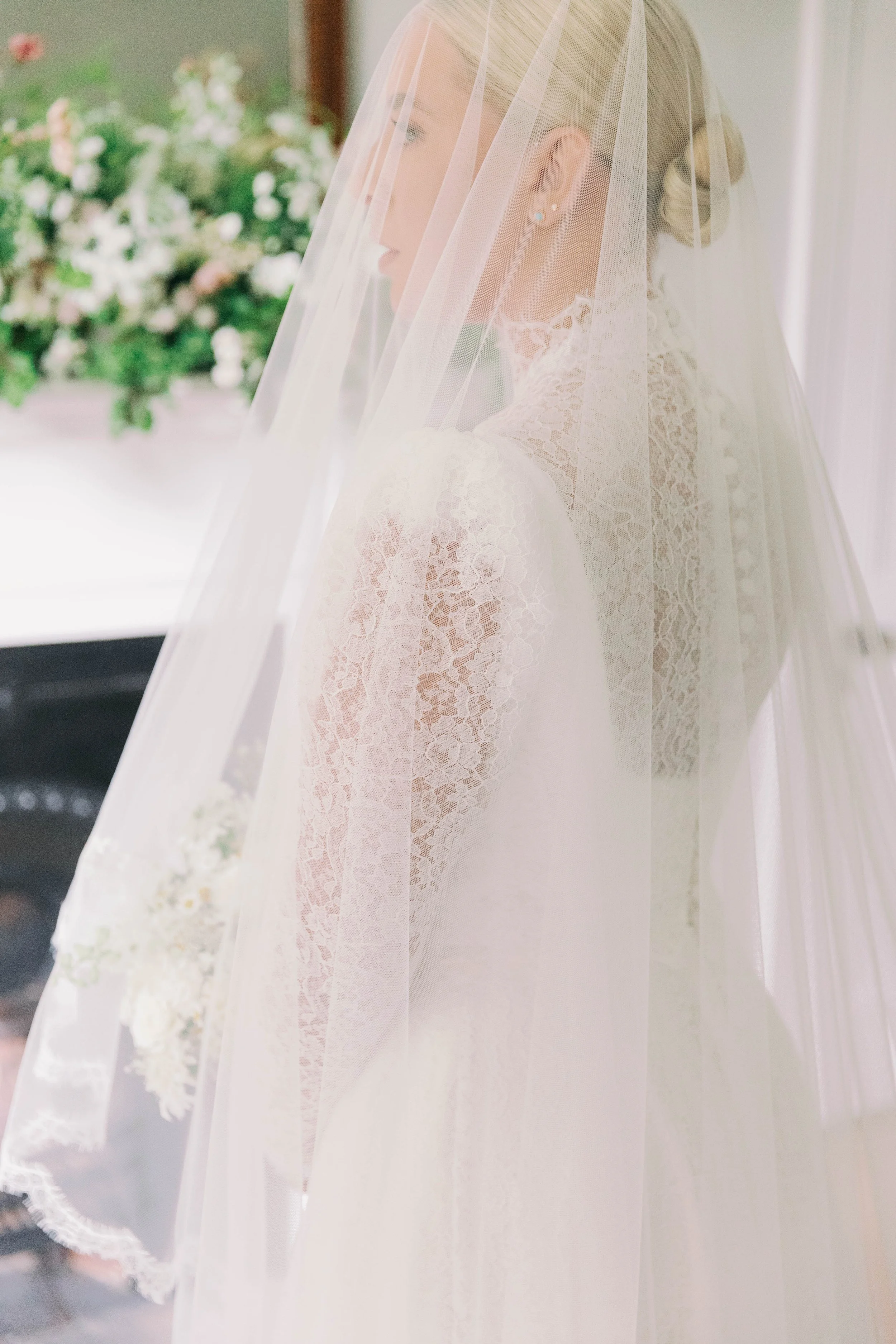 A bride wearing a white lace wedding dress and veil, standing near a window, with a blurred flower arrangement on a fireplace in the background. Private home wedding in Norfolk