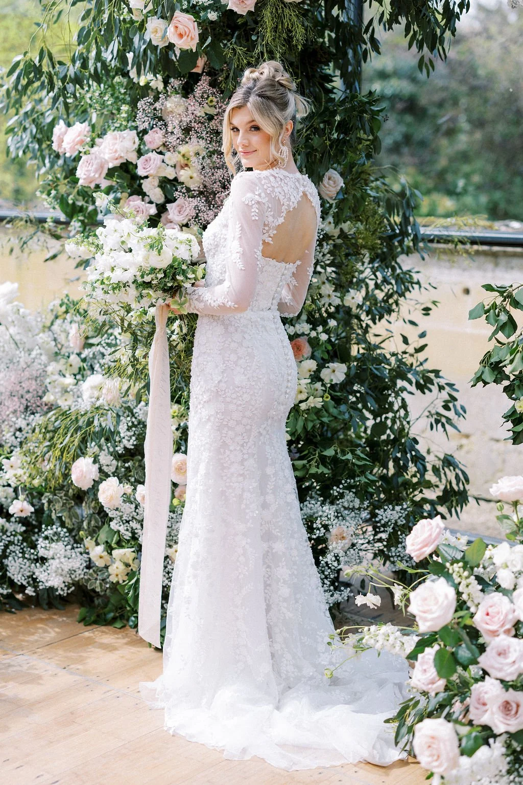 A bride in a white lace wedding gown holding a bouquet of white and green flowers, standing in front of a lush floral backdrop of pink, white, and green flowers and greenery.