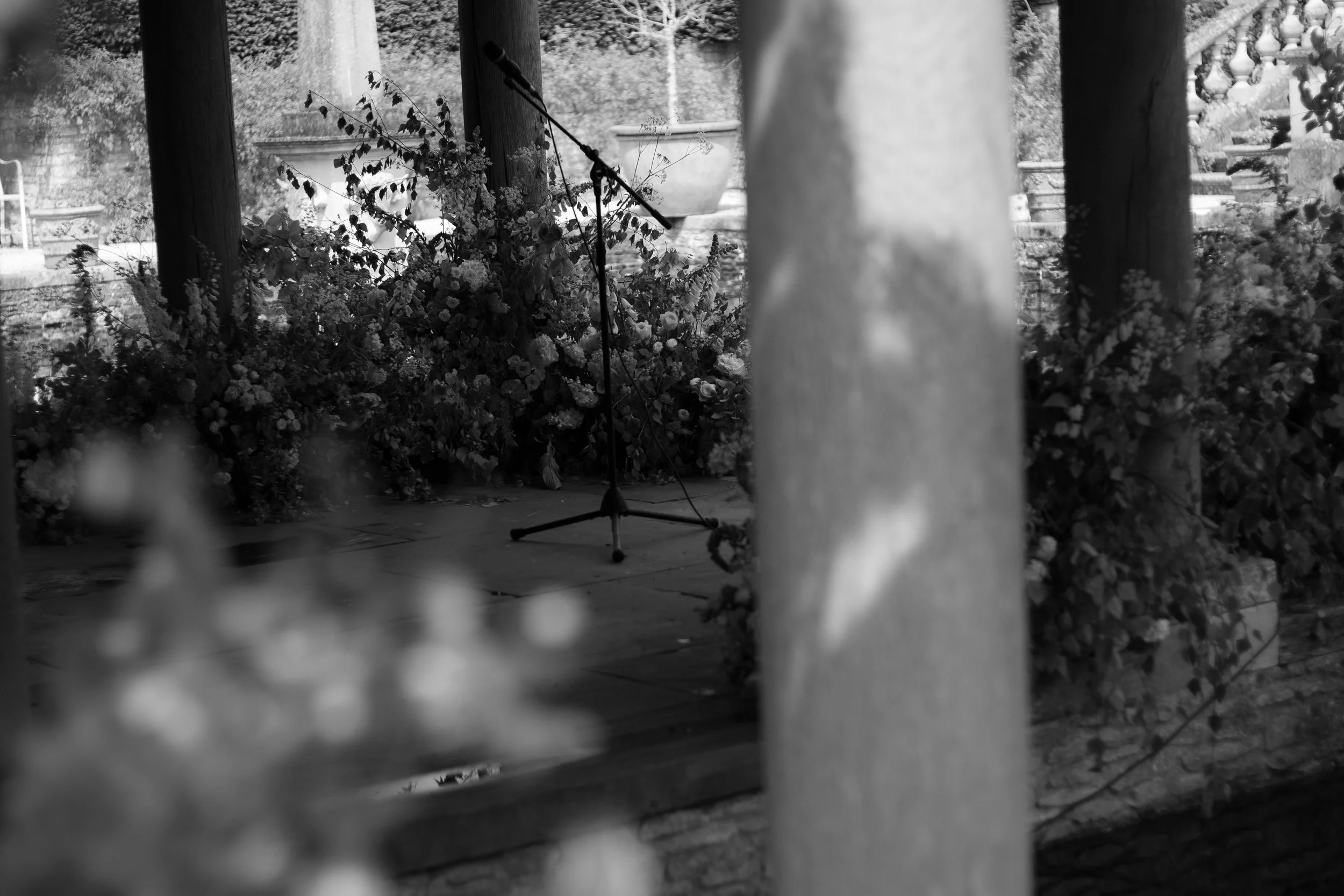Black and white photo of a microphone stand on a stage with floral arrangements in the background and a large stone urn in the distance.