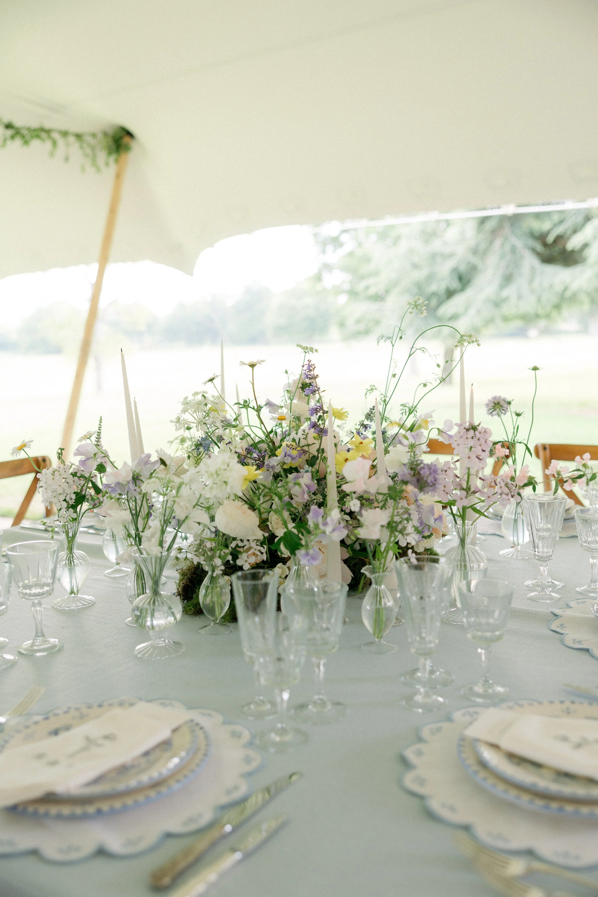Table centerpiece with an assortment of white and pastel-colored flowers in glass vases, set for a formal event outdoors under a large white tent.