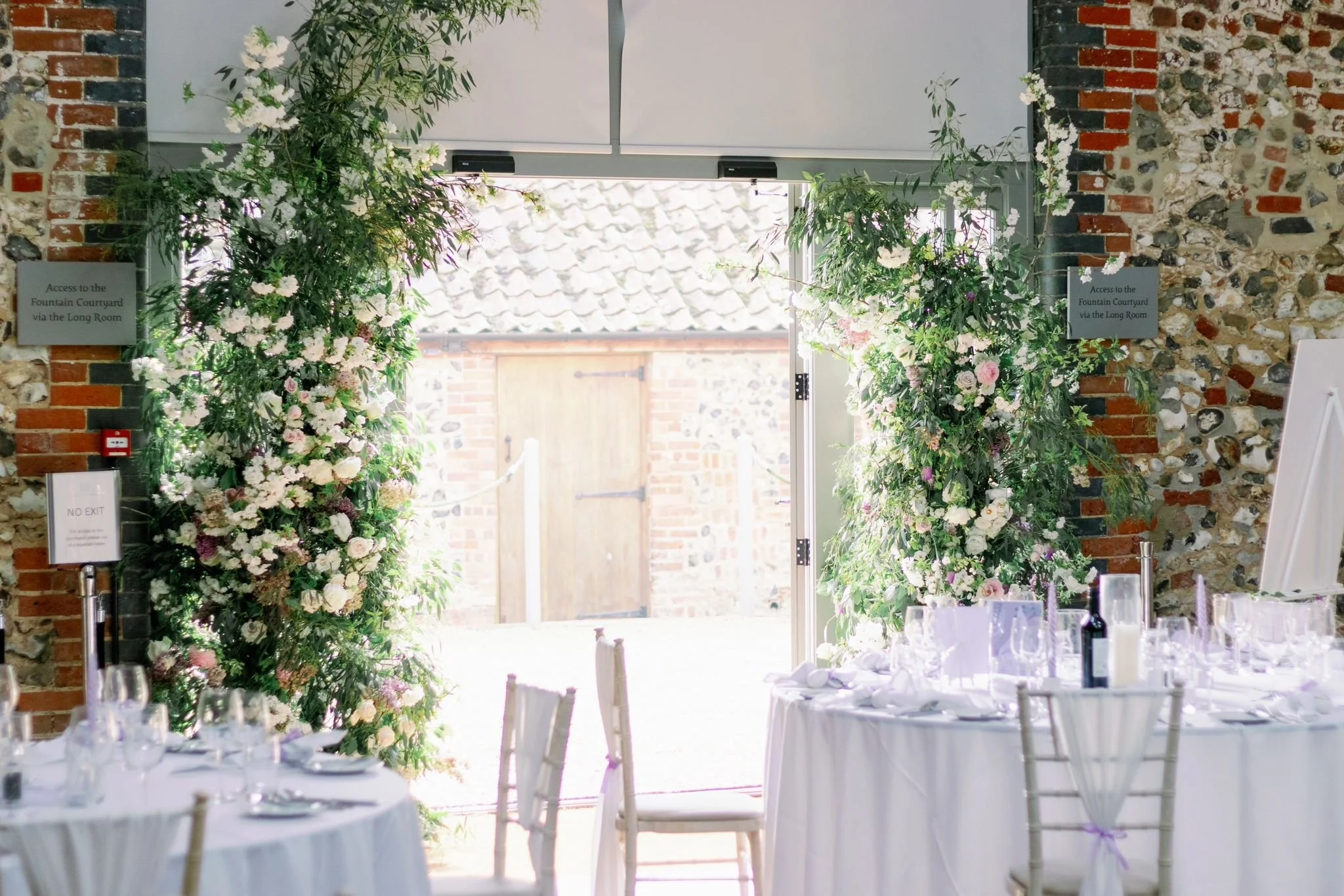 Decorated wedding or banquet reception area with white tables, chairs, and tall floral arrangements near open doors leading outside to a brick paving pathway.