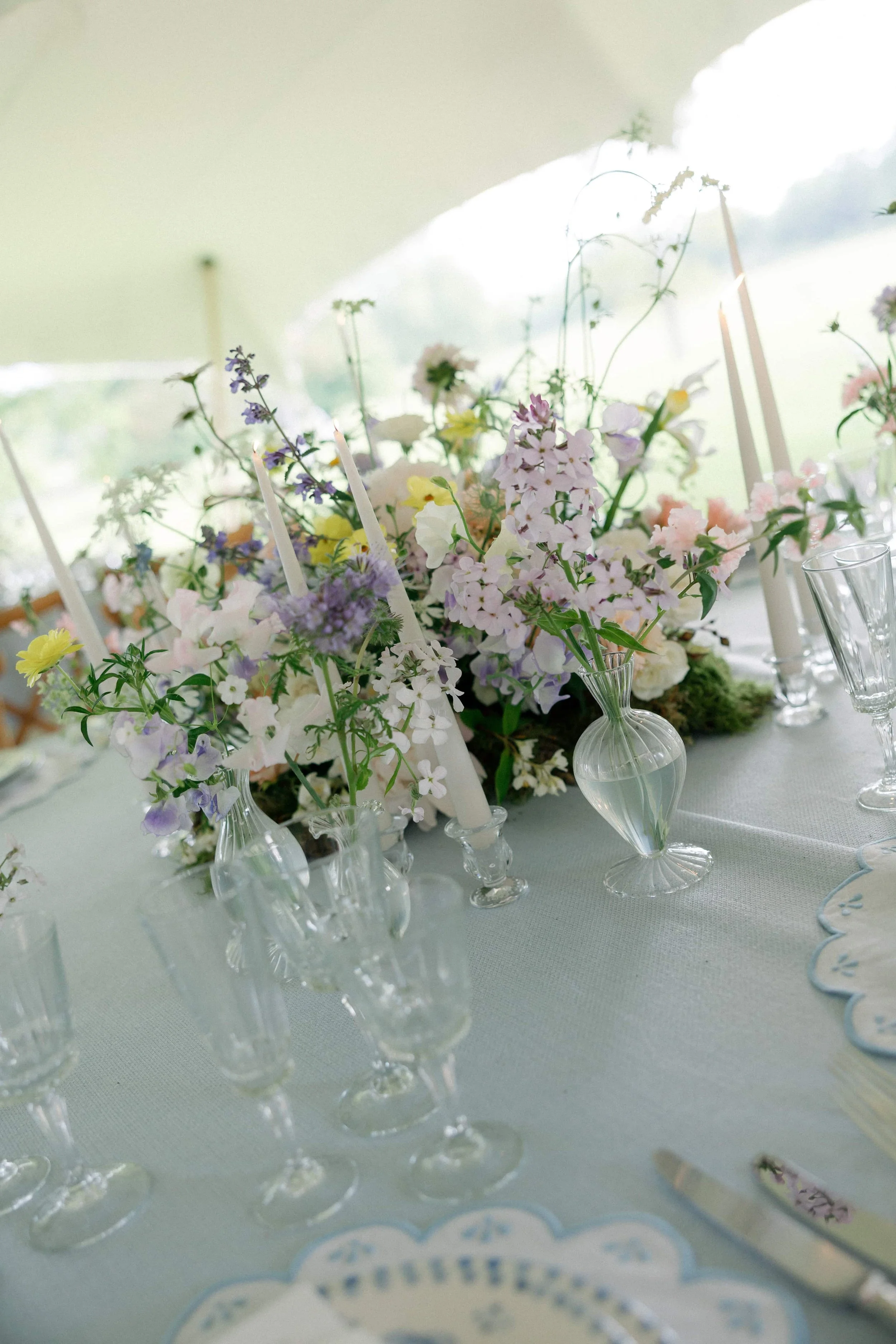 A table decorated with a floral centerpiece, vases, candles, and glassware at a wedding or event. Wilderness Reserve, Suffolk Florals By The Botany House