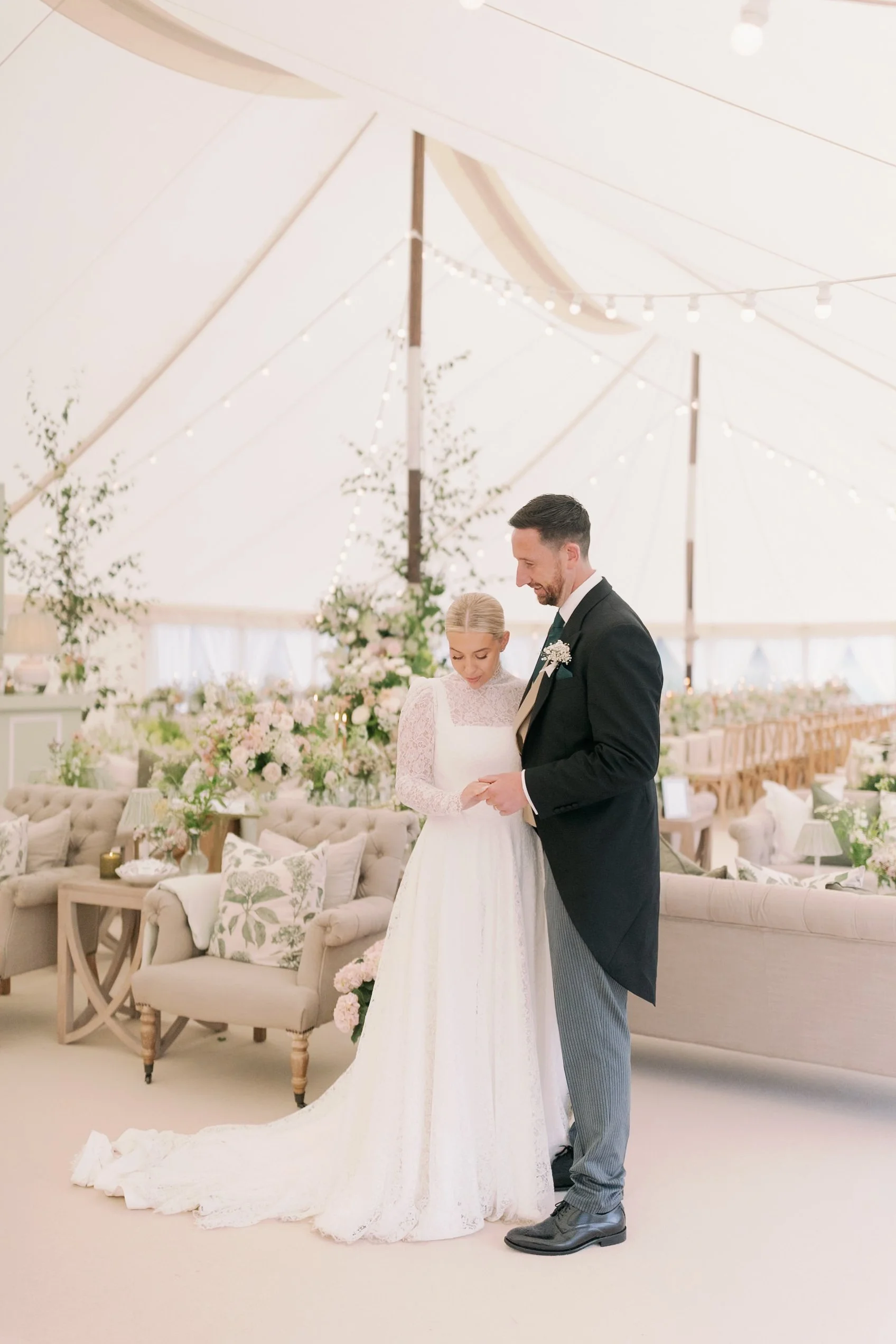A bride and groom standing together in a decorated wedding marquee, looking at something in the bride's hands, surrounded by floral arrangements and romantic lighting in Norfolk
