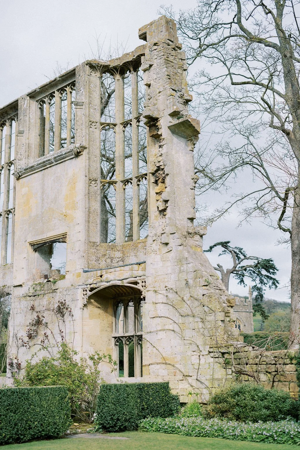 Ruined stone building with Gothic windows, partially collapsed, surrounded by bushes and trees.