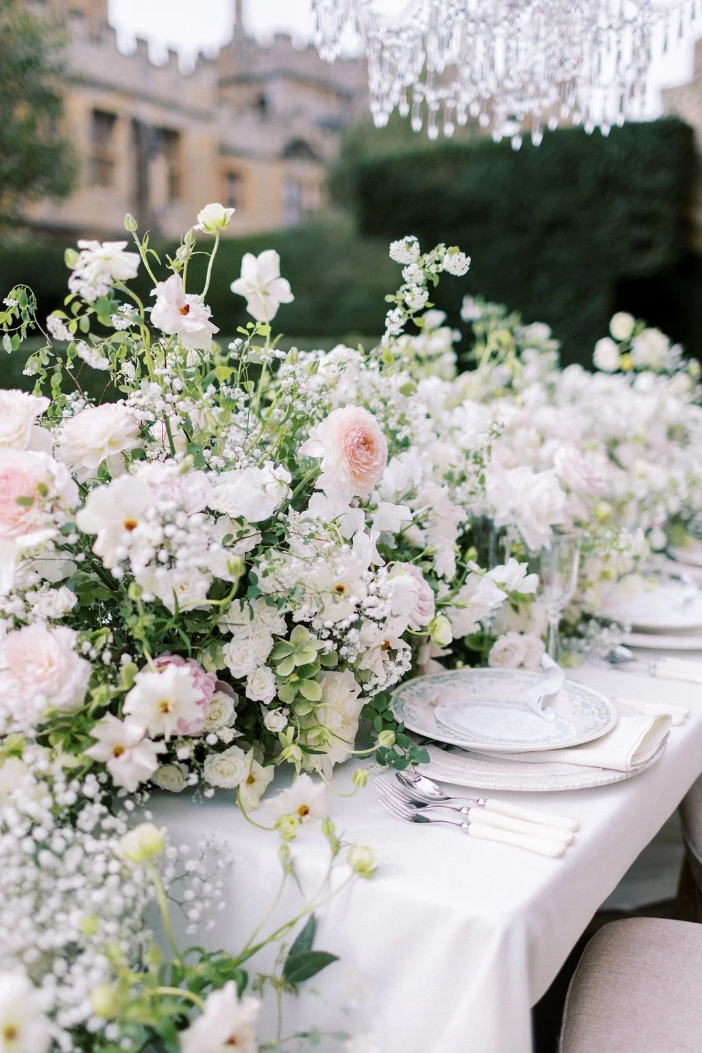 Elegant outdoor event table decorated with a large arrangement of white and light pink flowers, with china, silverware, and cream-colored napkins set on a white tablecloth.