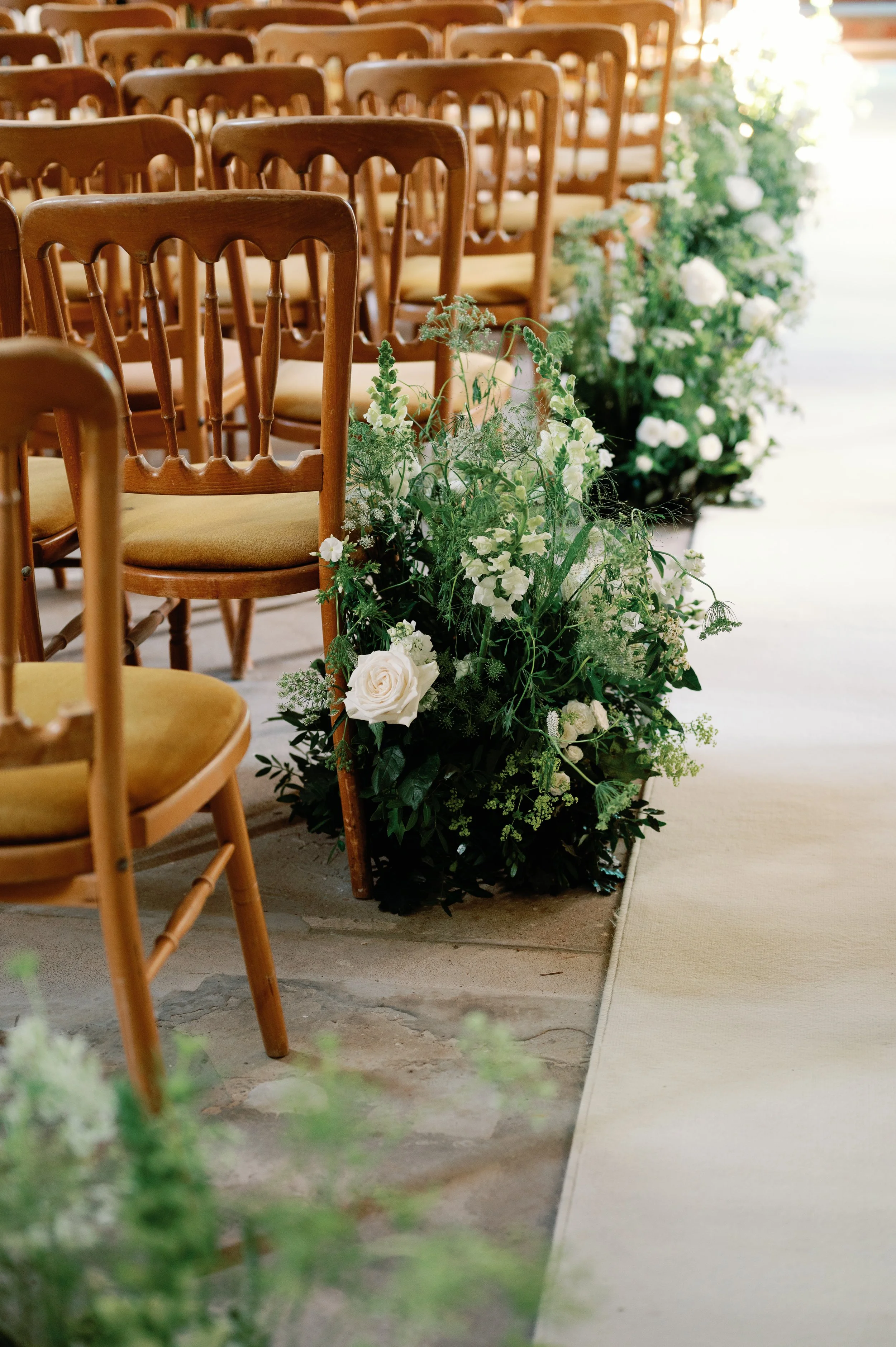 Wooden chairs with yellow cushions arranged in rows, decorated with white flowers and greenery along the aisle in a ceremony setting.