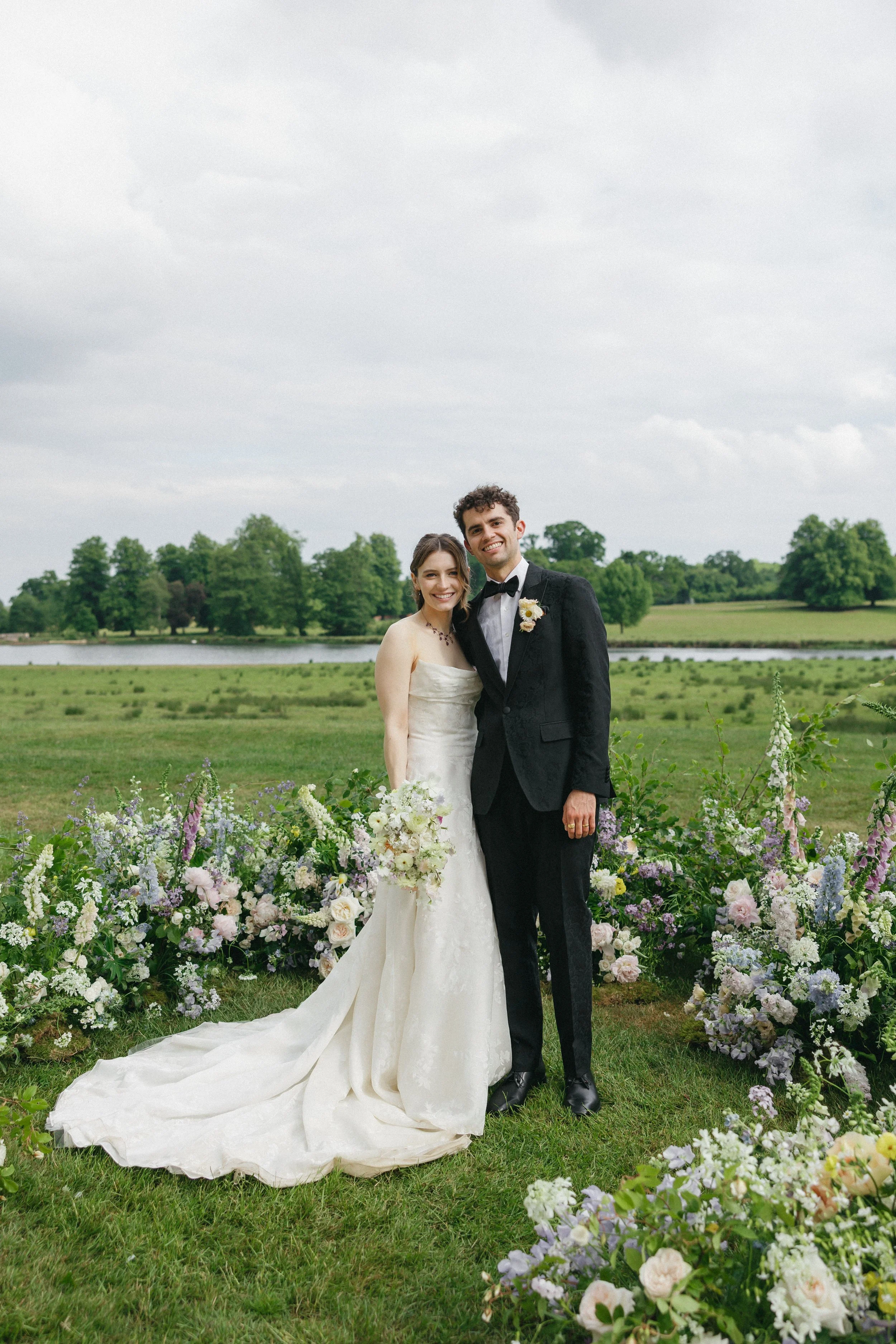 A bride and groom standing outdoors on a wedding day, smiling and holding a bouquet, surrounded by a floral arrangement with greenery and pastel-colored flowers, with a lake and trees in the background.