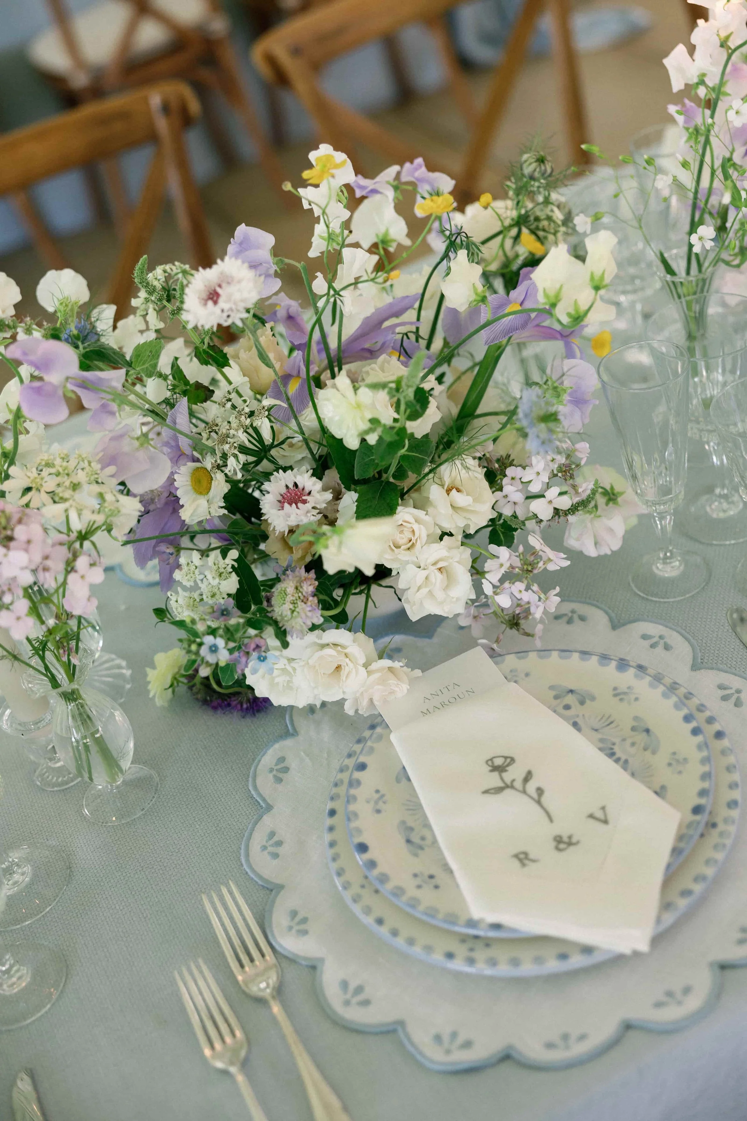 Elegant table setting with a floral centerpiece , a patterned blue and white plate, a monogrammed napkin, and clear glassware, in a decorated dining area. The Botany House Florist at Wilderness Reserve, Planned by Attabara Studio