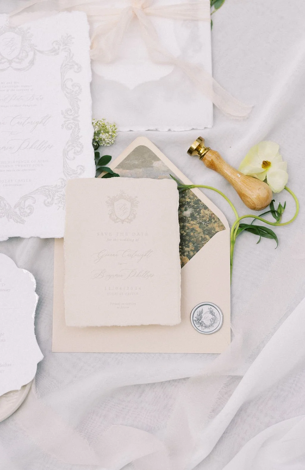 Elegant wedding invitation set with textured cream and white papers, a wax seal, a bouquet stamp, greenery, a brayer, and a cream flower on a white fabric background.