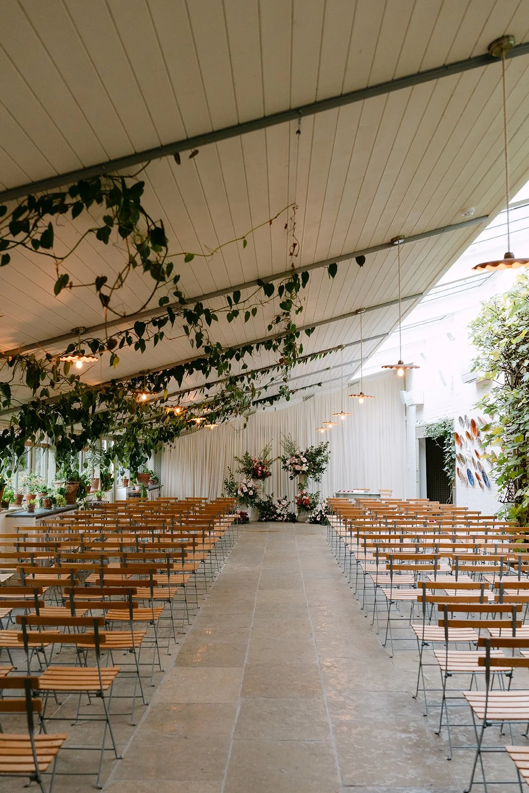 Indoor wedding ceremony setup with wooden chairs arranged in rows on either side of a central aisle, decorated with floral arrangements at the front, and greenery hanging from the ceiling. Kin House The Cotswolds The Botany House Florist