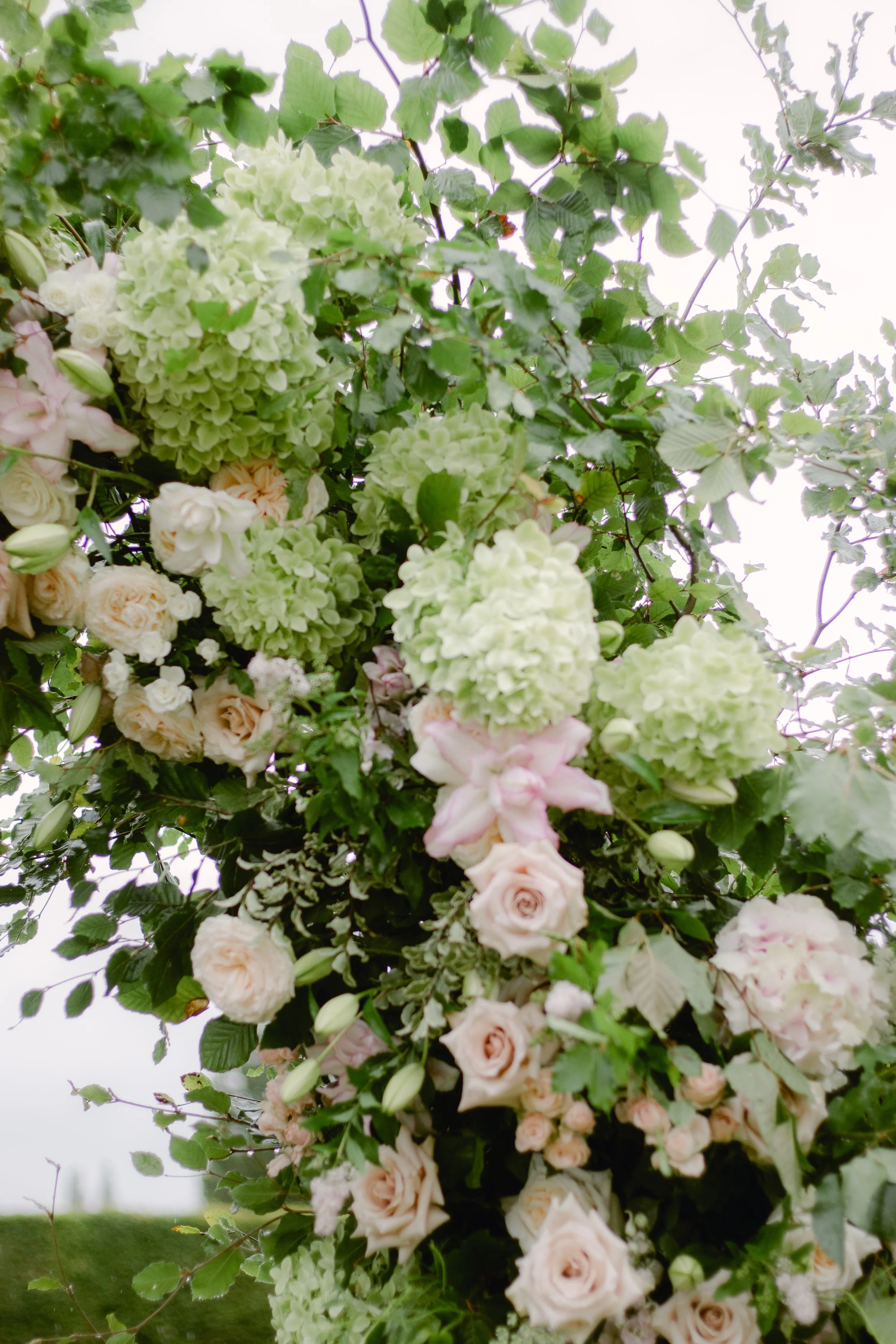 A close up of a floral archway with pale pink roses, light green hydrangeas, lilys, and other flowers with green leaves.
