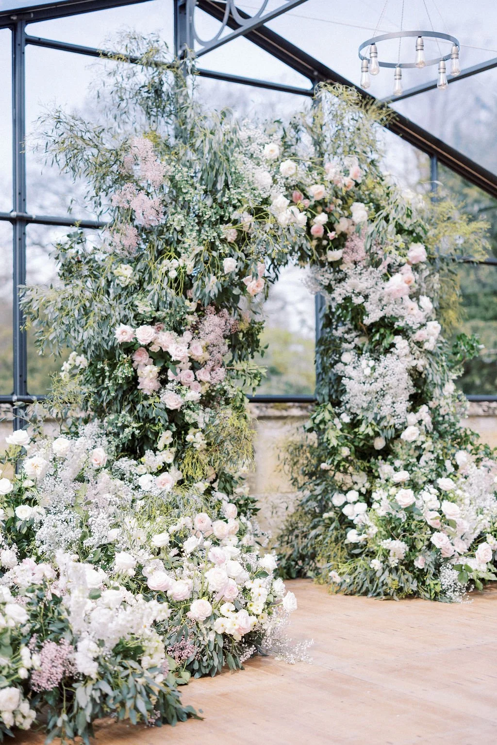 Floral arch decorated with white and light pink flowers and green foliage inside a glass-walled venue.