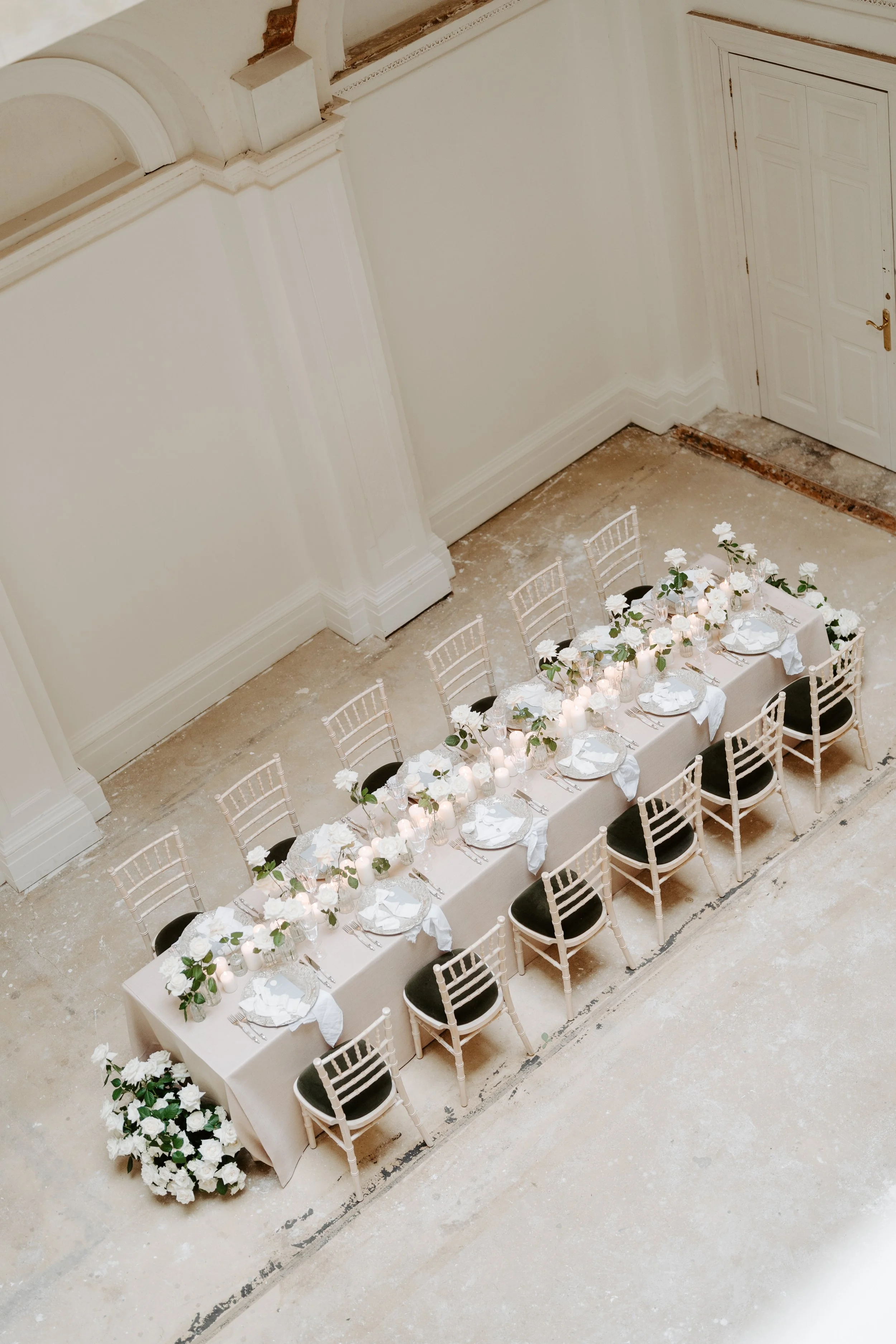 Long table decorated with white flowers, candles, and place settings, arranged for a formal event, in a room with high ceilings and a white door.