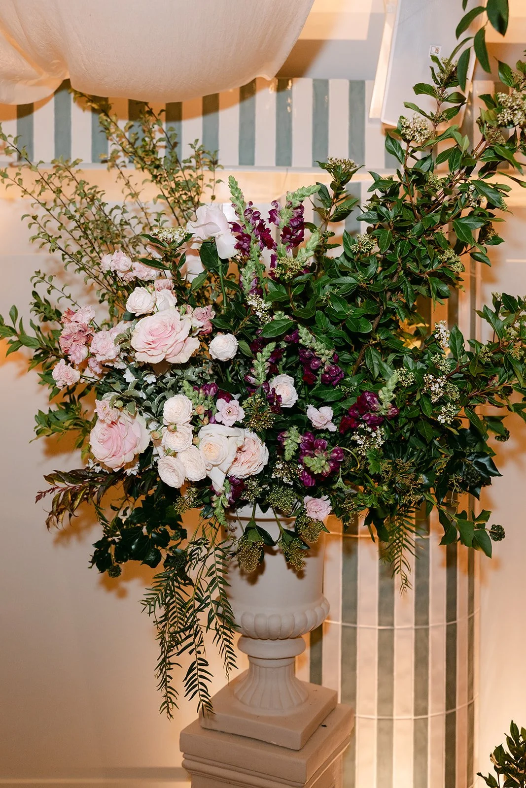 A large elegant floral arrangement in a classical white urn with pink, white, and purple flowers, surrounded by greenery, placed on a pedestal in a interior space. The Botany House Wedding Florist, Kin House The Cotswolds. Planned by Get wed