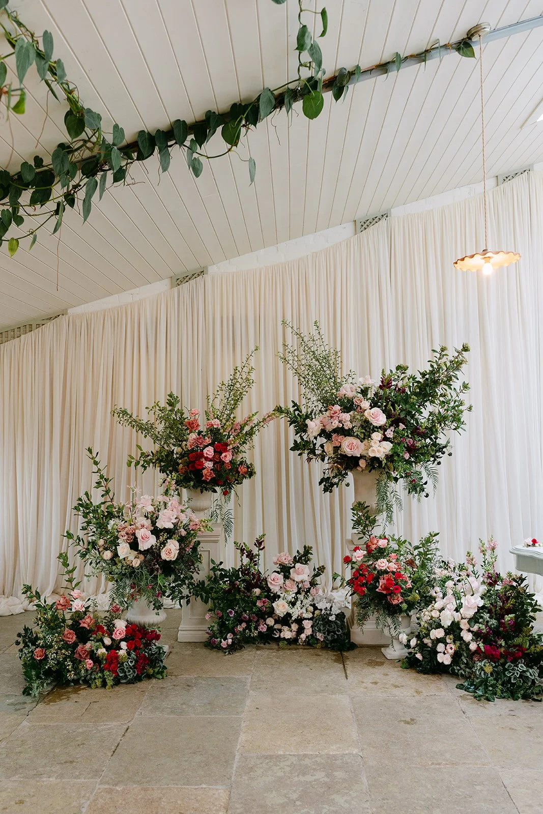 Beautiful indoor floral arrangement with various pink, white, and red flowers on white pedestals and the floor, set against cream draped curtains and a ceiling with white paneling and a hanging light fixture. Kin House The Cotswolds The Botany House