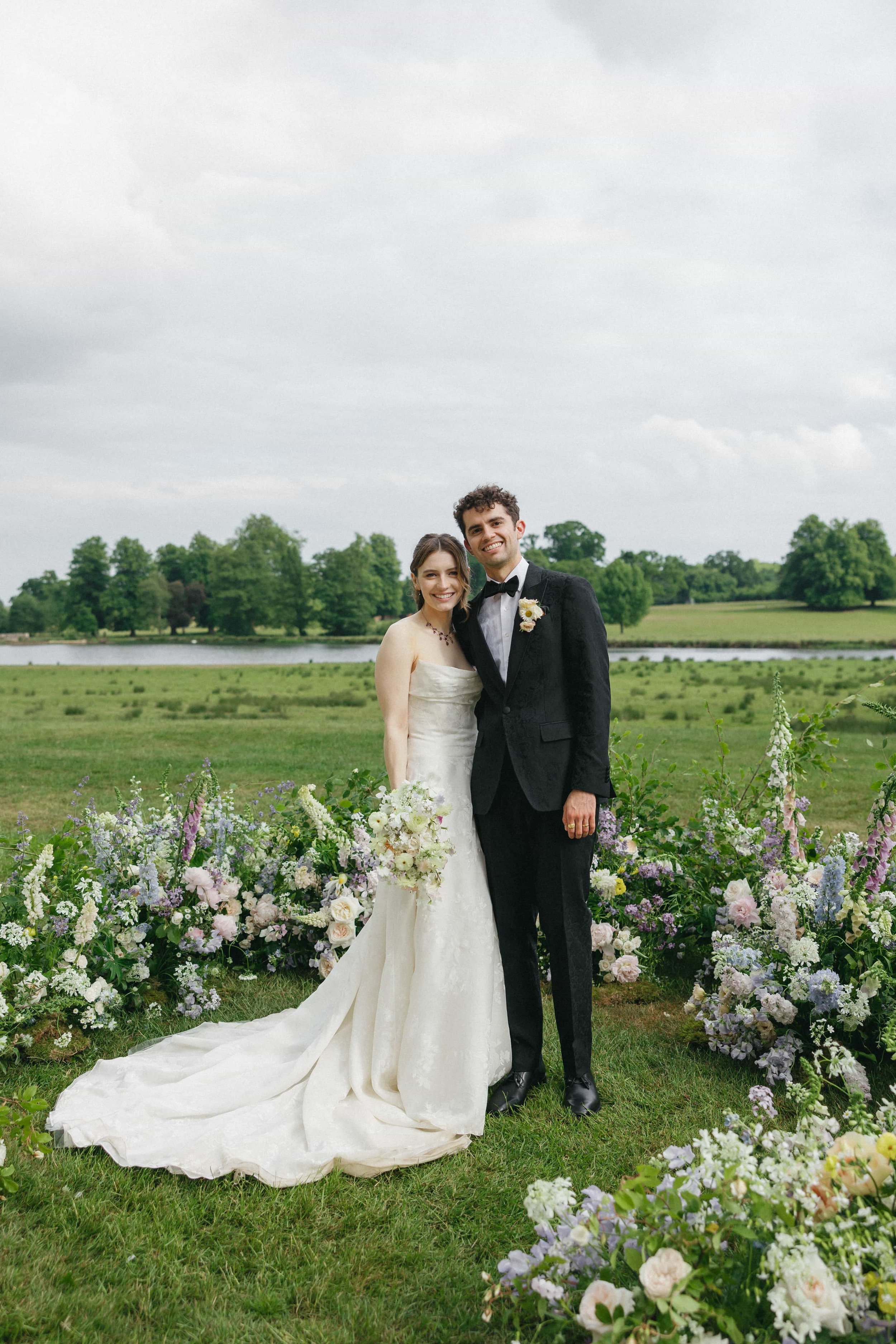 A bride and groom stand together outdoors on their wedding day, smiling, with a lush green landscape and a river in the background, surrounded by colorful flowers. Wilderness Reserve, Suffolk Florals By The Botany House