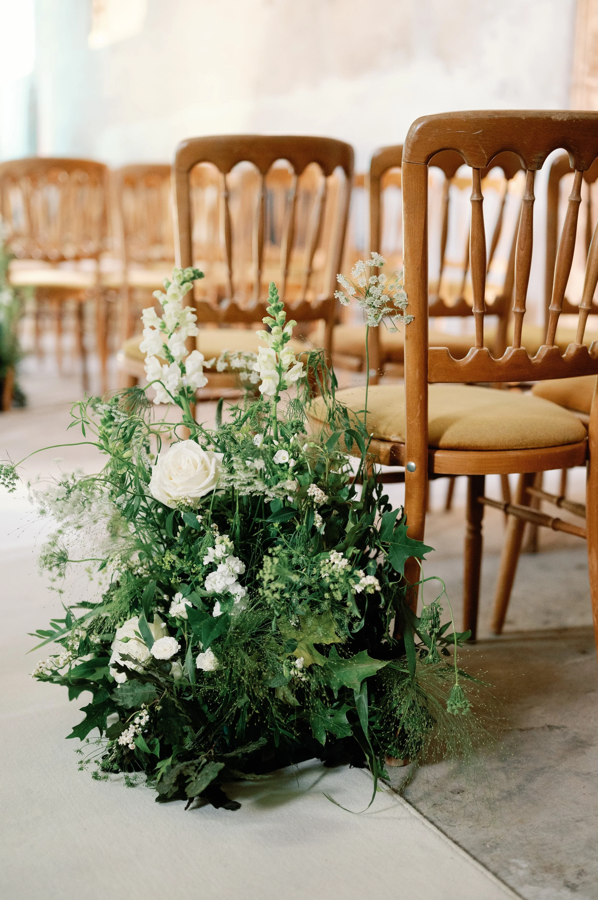 A floral arrangement with white flowers and greenery on the floor next to wooden chairs in a room with a concrete floor and a light-colored wall.