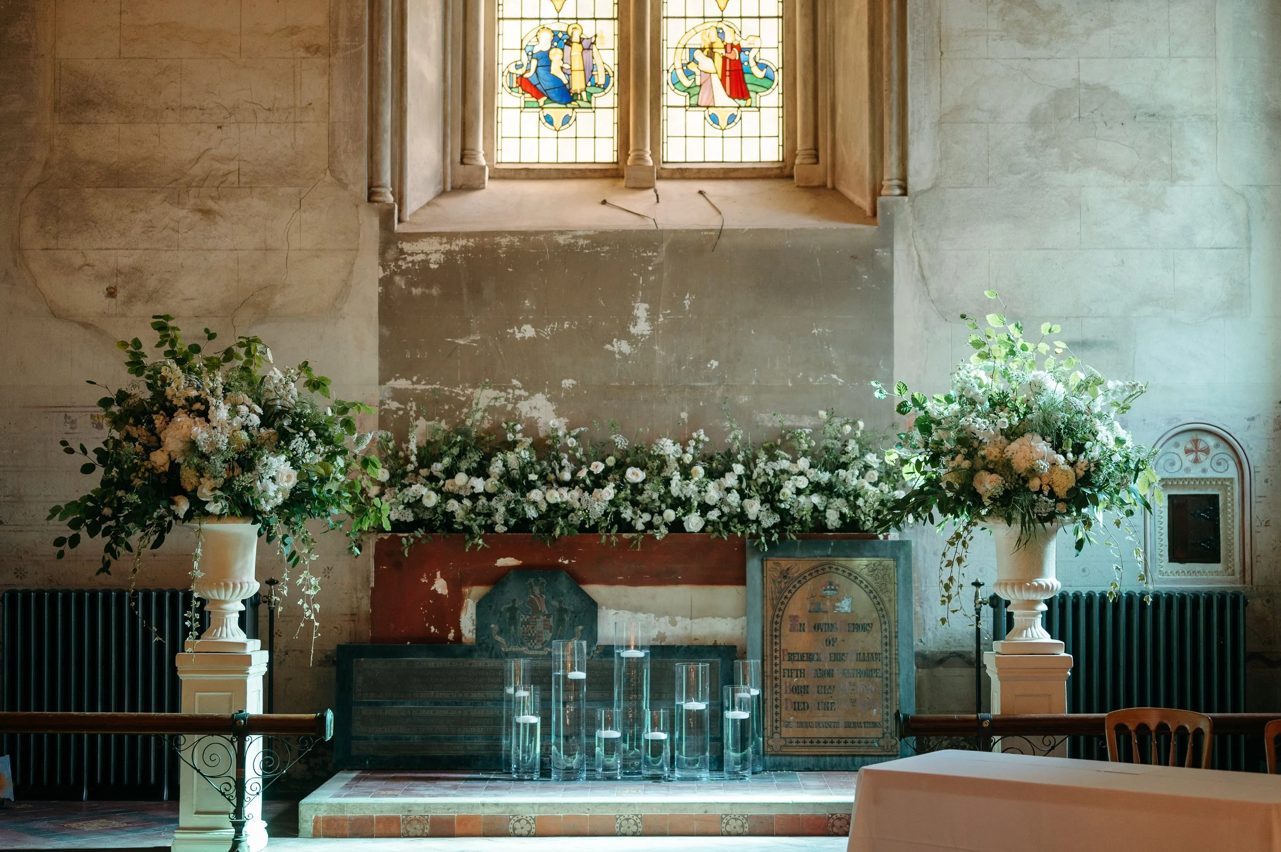 Interior of a church decorated for a wedding with white flowers, large glass vases with floating candles, stained glass windows, and an altar.