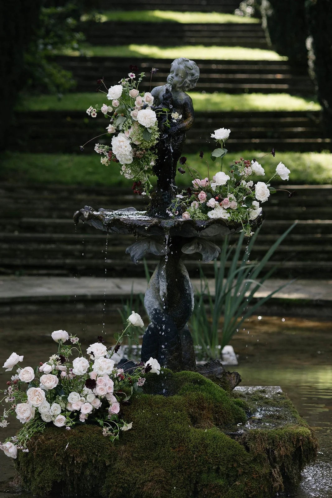 Decorative fountain with a cherub statue holding blush and cream flowers at Cornwell Manor, styled for a luxury garden-style wedding by The Botany House, best luxury wedding florist.