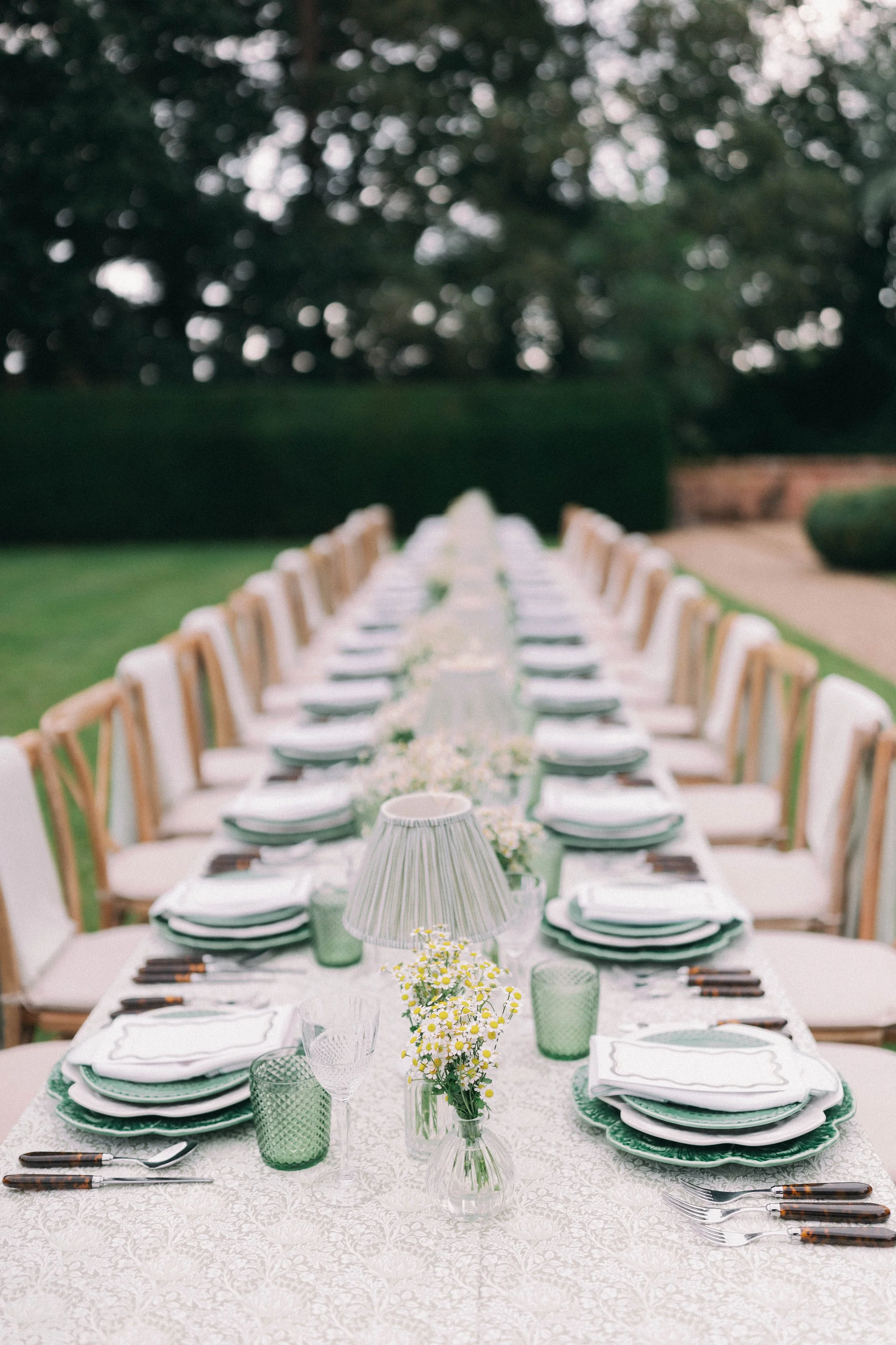 Long outdoor dining table set with floral tablecloth, green glassware, white plates with green trim, floral centerpieces, table lamps, and coordinating chairs, in a garden setting in Norfolk